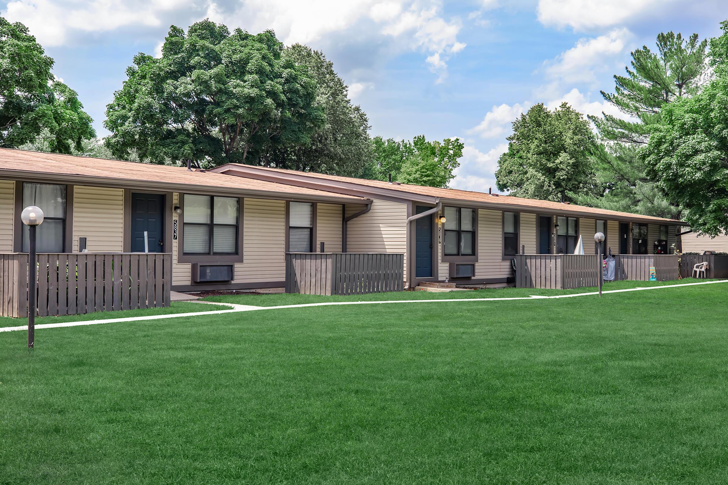 A row of single-story apartment units with light-colored siding, surrounded by well-maintained green lawns and trees. Each unit has front steps and a small porch area, with streetlights positioned in front. The sky is partly cloudy, creating a bright and inviting atmosphere.