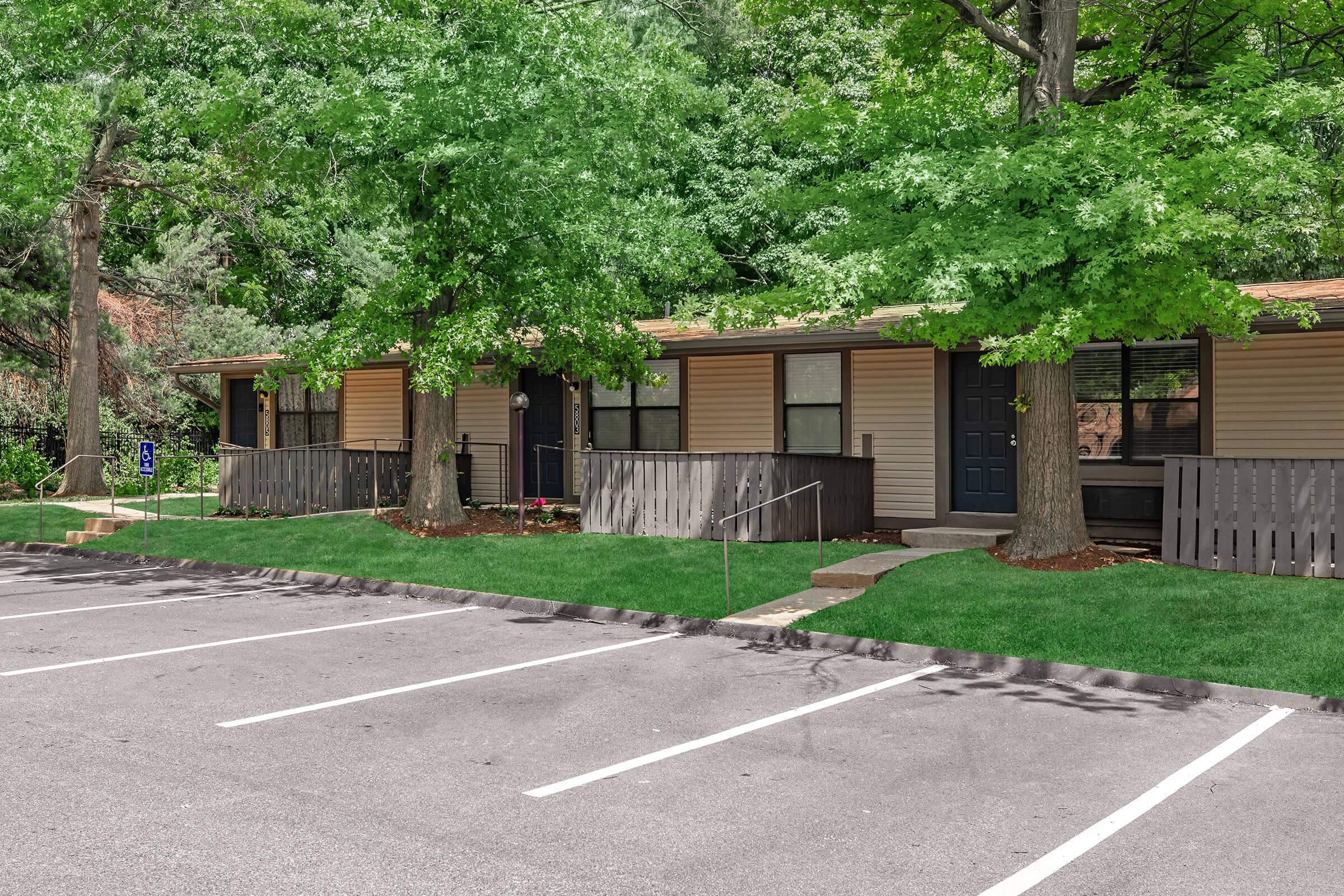 Row of residential buildings surrounded by trees, with a well-maintained green lawn in front. Parking spaces are visible in the foreground, and the walkway leads to individual entrances of the units. The atmosphere is peaceful and inviting, ideal for a suburban living environment.