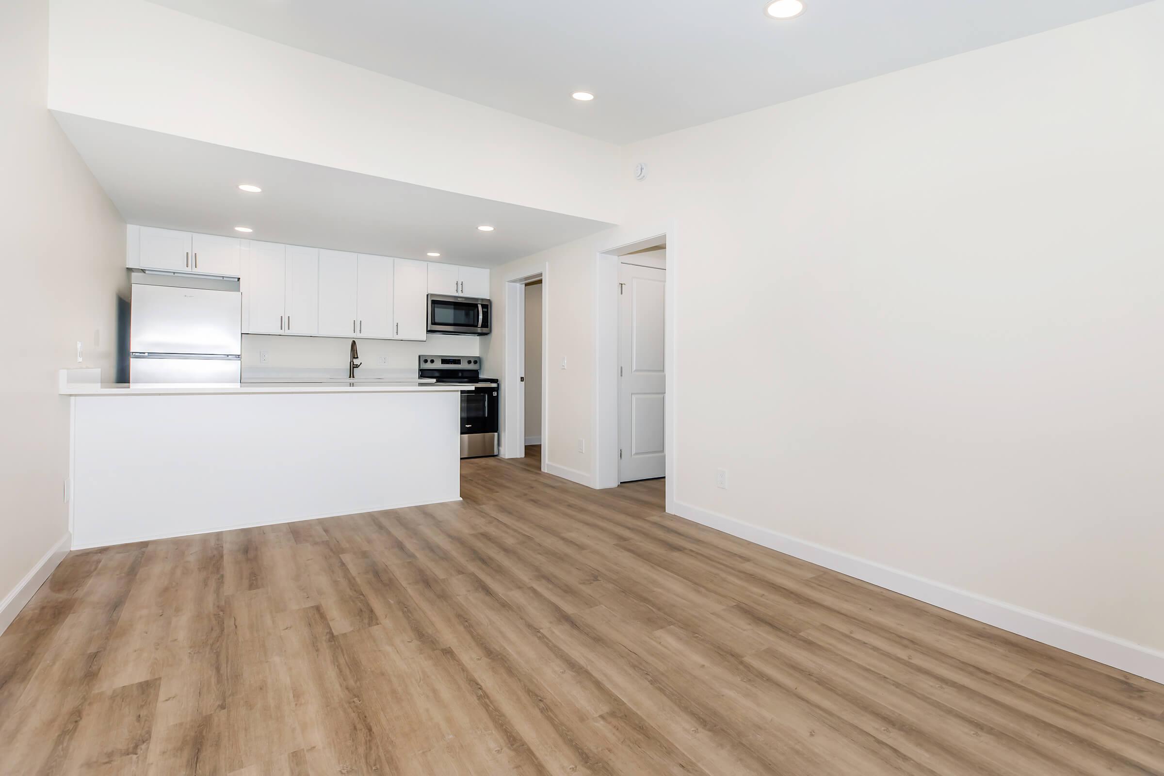 Modern, unfurnished kitchen and living area with light wood flooring. The kitchen features white cabinetry, stainless steel appliances, and a breakfast bar. The space is bright, with recessed lighting and an open layout leading to a doorway. Walls are painted in light, neutral colors.