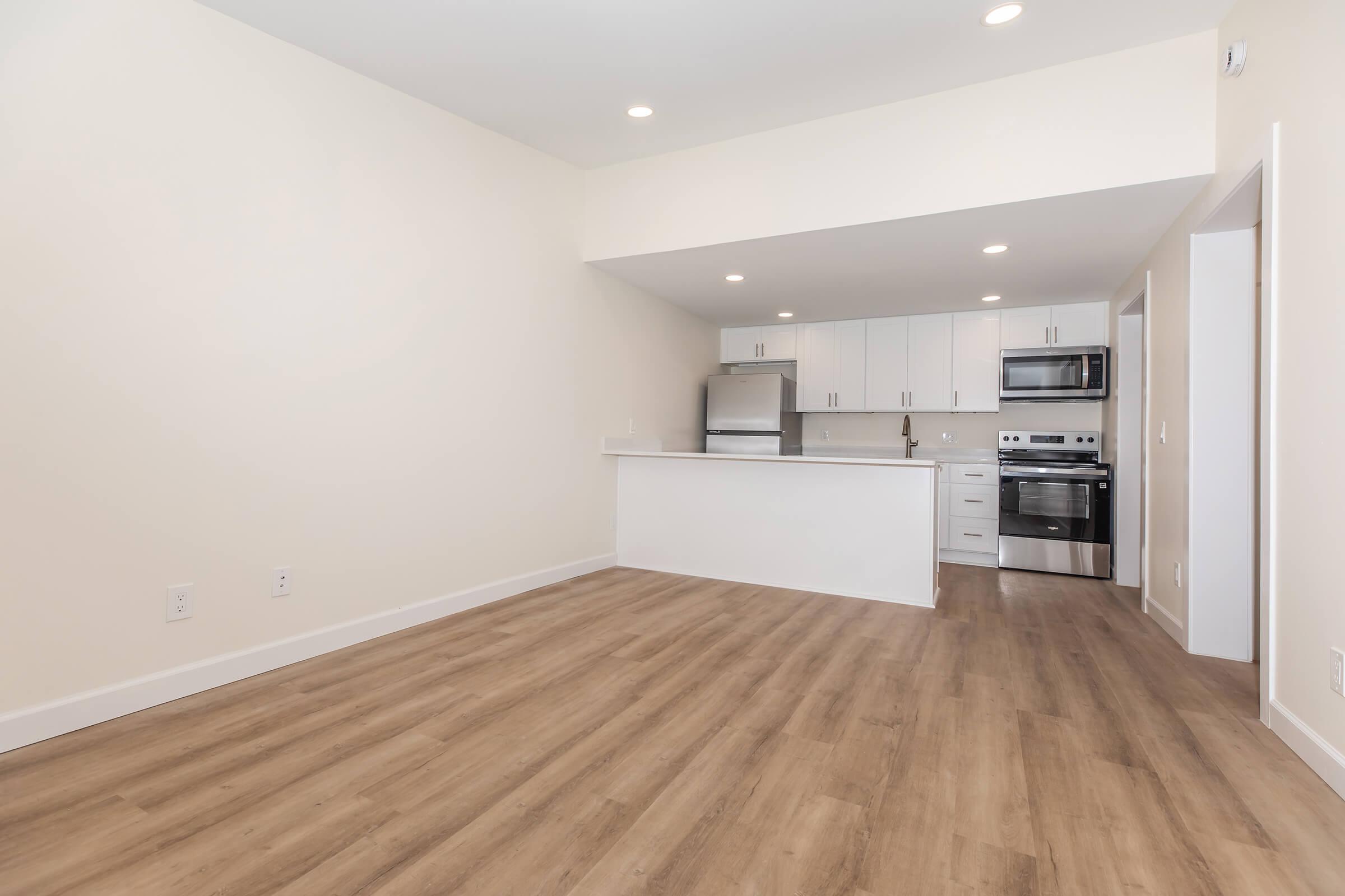 A spacious, modern kitchen and living area featuring light wood flooring, white cabinetry, stainless steel appliances, and bright overhead lighting. The room has neutral-colored walls, creating a clean and inviting atmosphere.