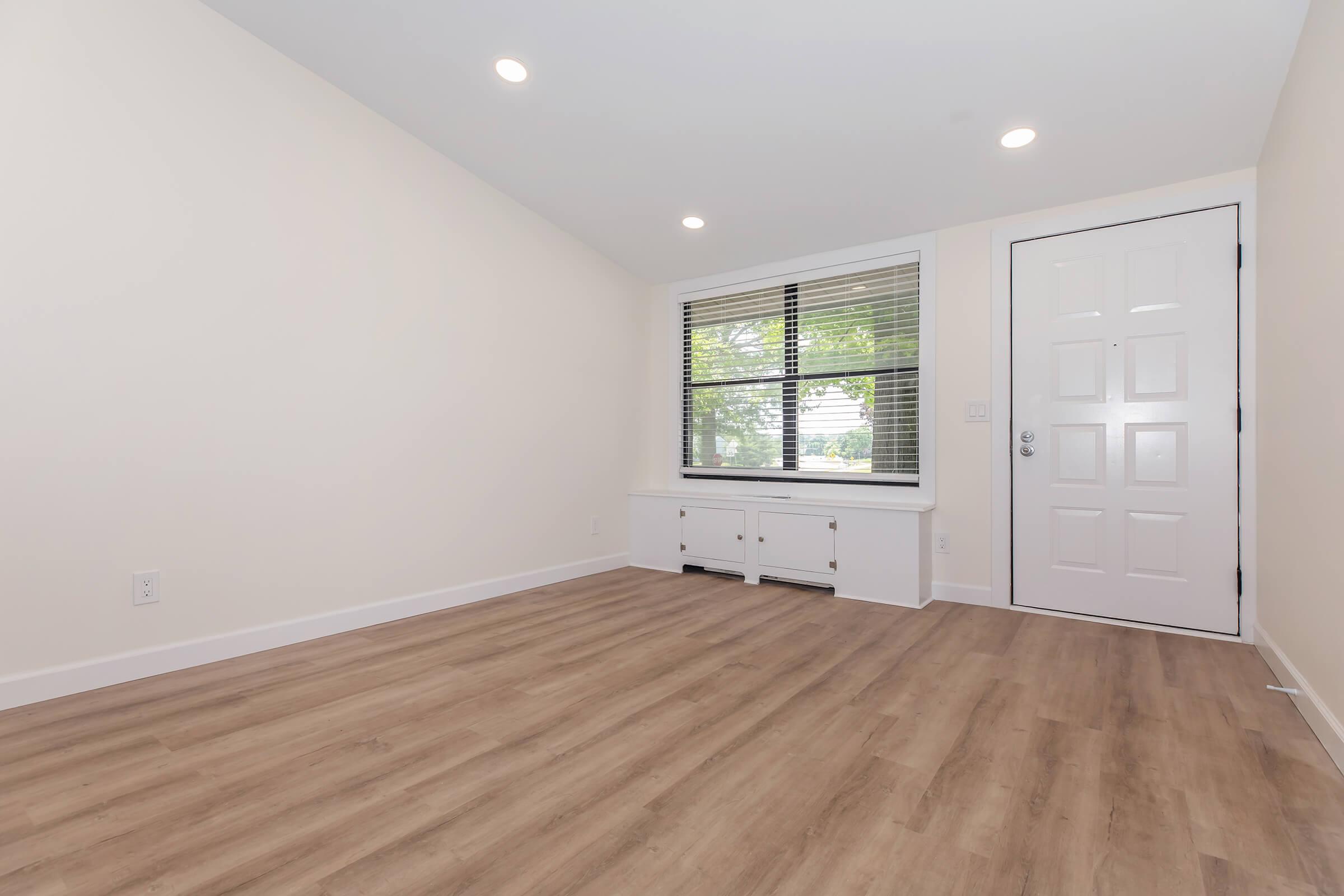 A bright and modern interior space featuring light-colored walls, a large window with blinds, and a white door. The floor is covered with light wood laminate. There are built-in cabinets beneath the window, creating a clean and spacious atmosphere, ideal for various uses such as a living room or office.
