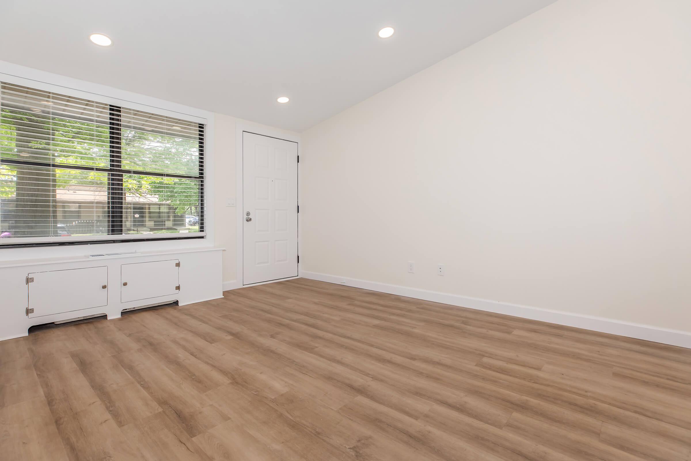 A spacious, empty room with light-colored walls and wooden flooring. It features a large window letting in natural light, a white door, and a built-in white cabinet beneath the window. The overall atmosphere is bright and inviting, perfect for personal decoration.