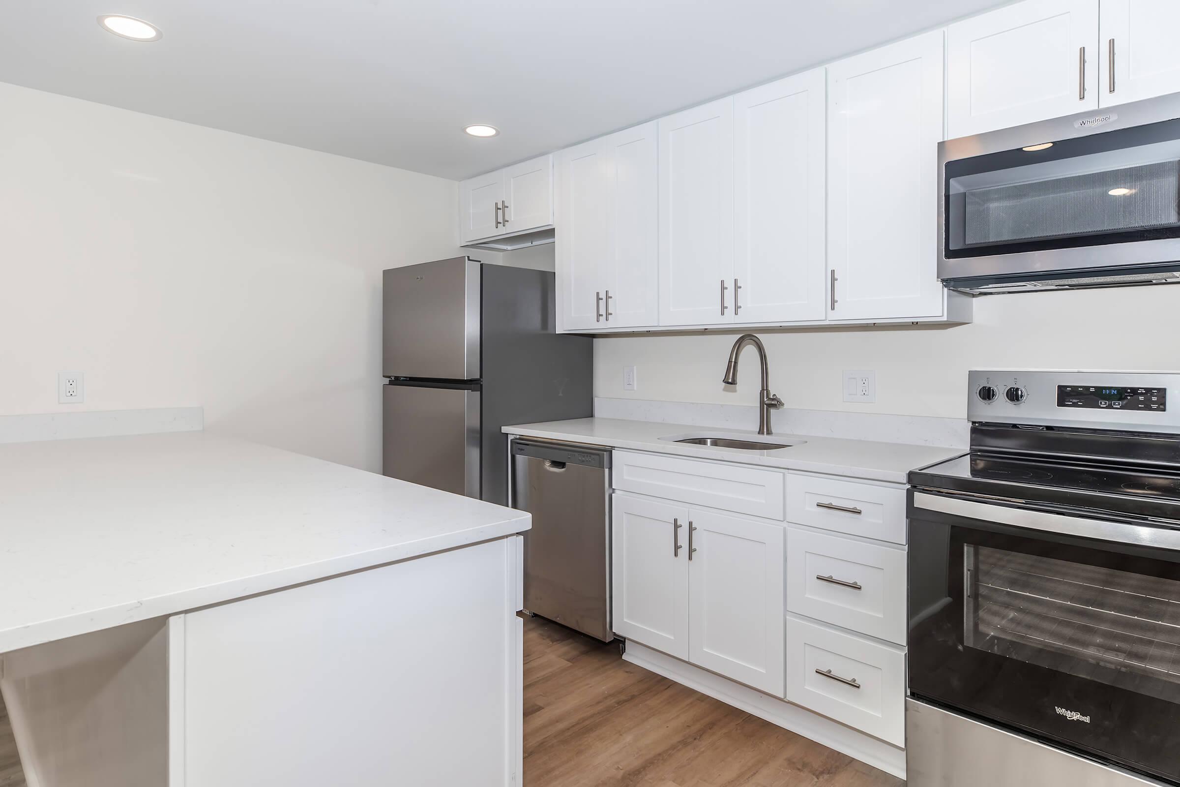 Modern kitchen with white cabinetry, stainless steel appliances including a refrigerator, microwave, and oven. The countertop is light-colored with a clean design, and there's a sink with a contemporary faucet. The space features natural wood flooring and recessed lighting for an inviting atmosphere.