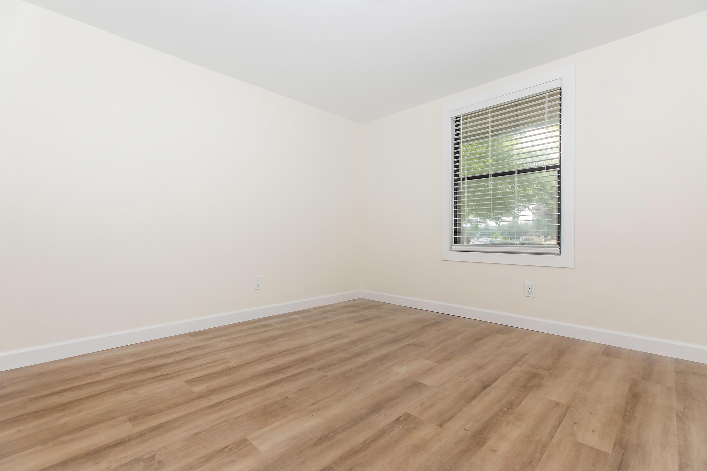 Empty room with light-colored walls and wooden flooring. A window with blinds is visible on the right side, allowing natural light to enter. The space appears clean and minimalistic, suitable for various uses or furnishings.