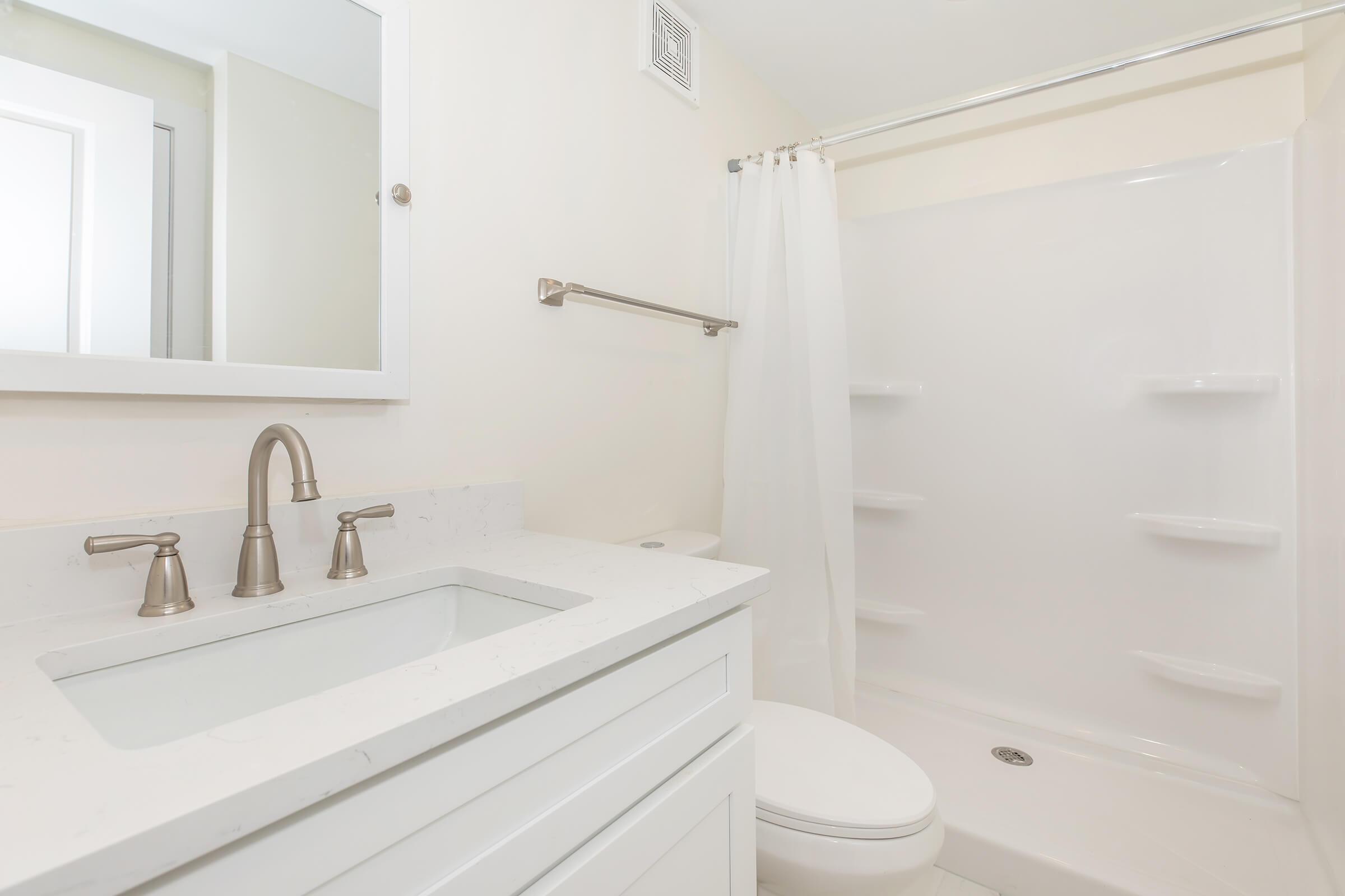 A modern bathroom featuring a white vanity with a rectangular sink and brushed nickel faucets, a large wall mirror, a shower area with a clear curtain, and a light-colored tile floor. The walls are painted in a soft beige, creating a bright and clean aesthetic.