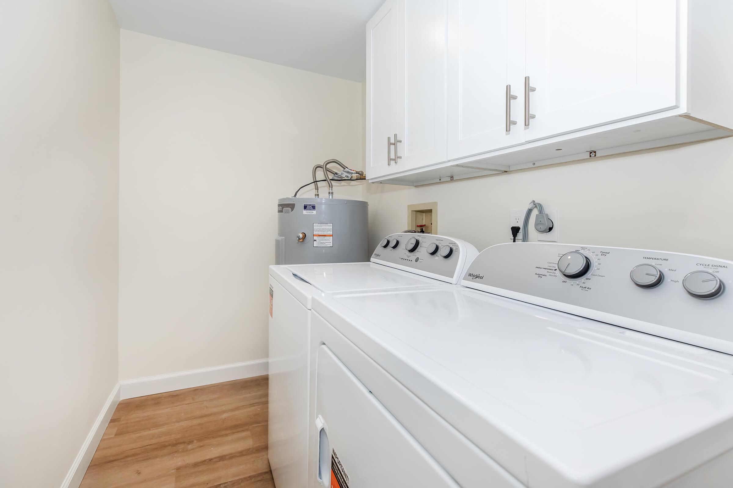 A clean laundry room featuring a white washing machine and dryer side by side. Above them is a shelf with closed white cabinets. In the background, there is a gray water heater. The floor is light wood laminate, and the walls are painted a soft beige.