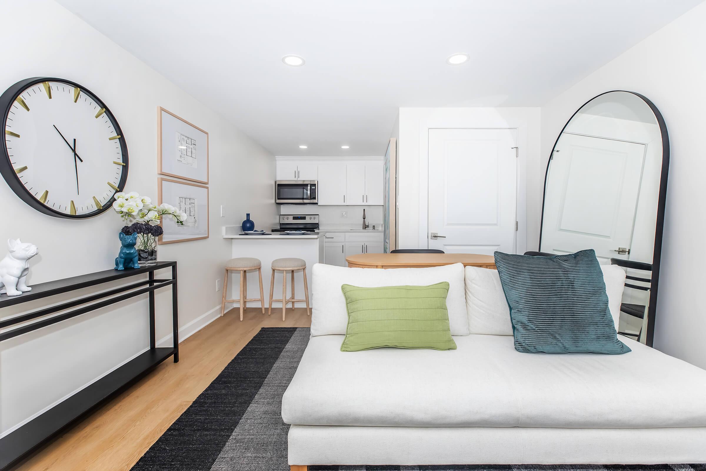 Modern living space featuring a light-colored sofa with green and blue accent pillows. A large mirror reflects the room, which also includes a wall clock, framed artwork, and a decorative shelf with vases. The kitchen area is visible in the background, equipped with modern appliances.
