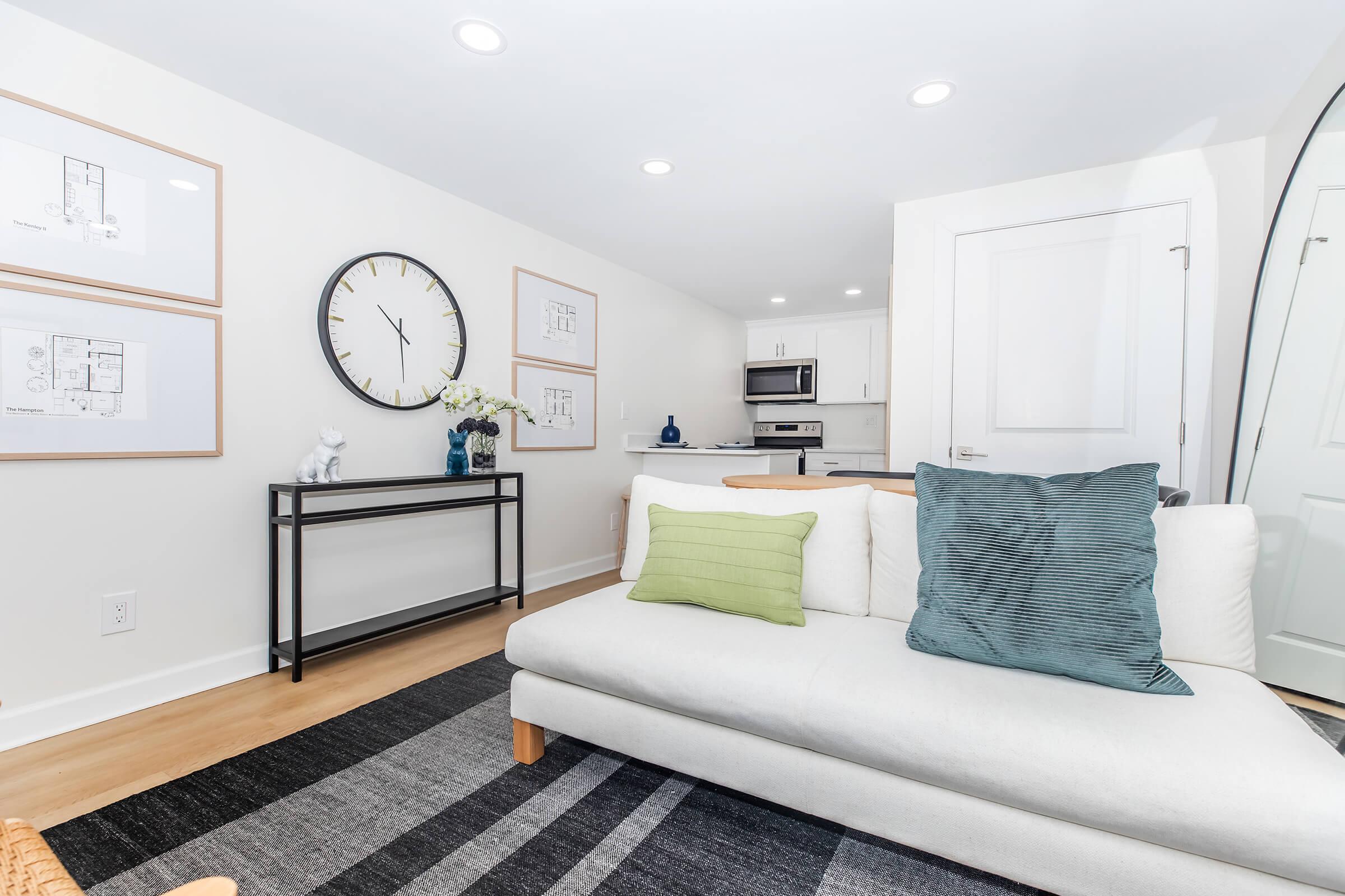 A modern living room featuring a white sofa with green and blue cushions, a black console table with decorative items, a large wall clock, framed artwork, and a kitchen area visible in the background. Natural light illuminates the space, creating a cozy and inviting atmosphere.