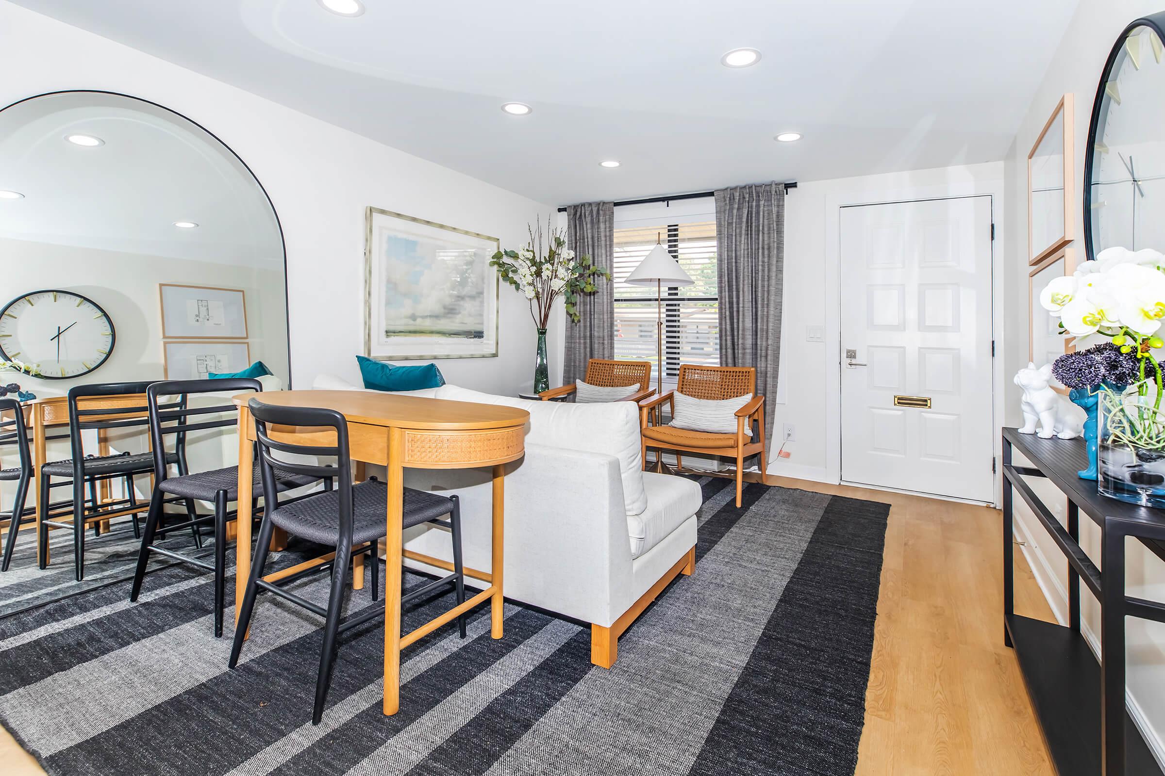 A modern living space featuring a dining table with chairs, two armchairs, and a decorative rug. The room is adorned with plants, artwork, and mirrors, with large windows letting in natural light. The front door is visible, adding to the cozy and inviting atmosphere of the area.