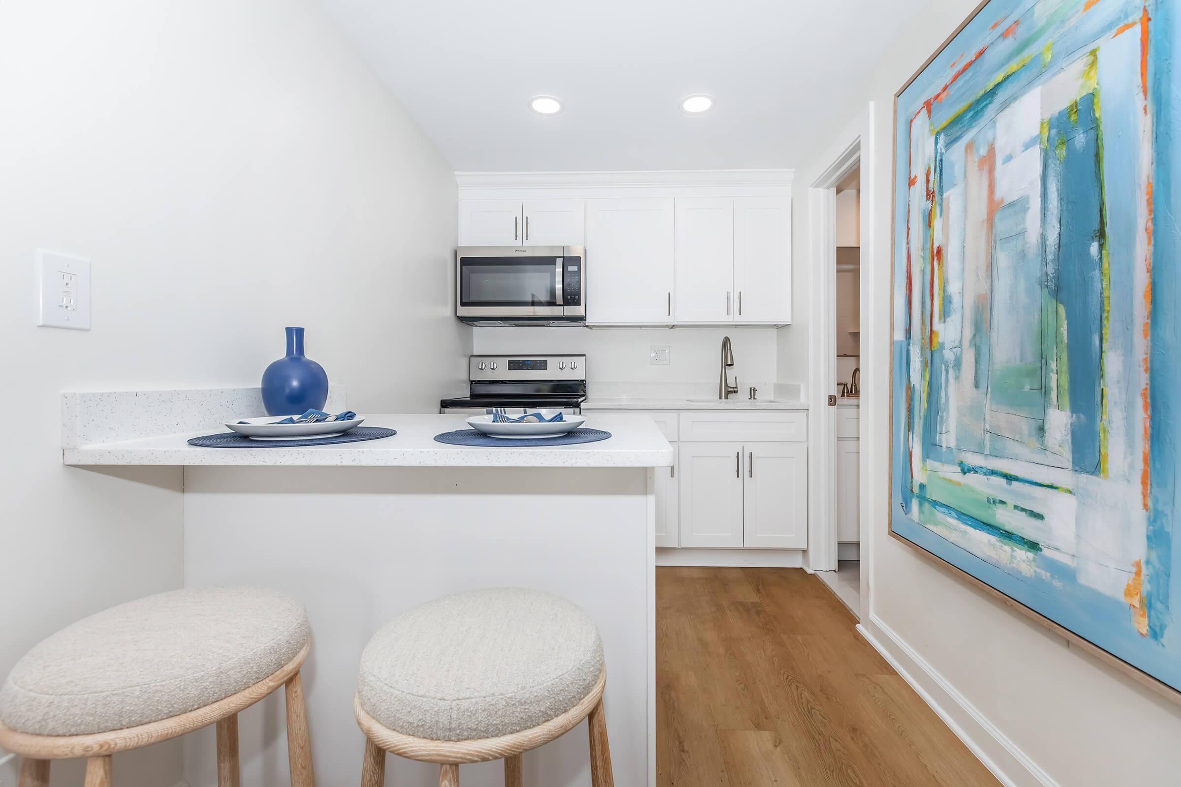 Modern kitchen featuring white cabinetry, stainless steel appliances, and a large colorful abstract painting on the wall. A small breakfast bar with two stools is visible, along with neatly set plates and a decorative blue vase. Bright lighting enhances the spacious and clean aesthetic.