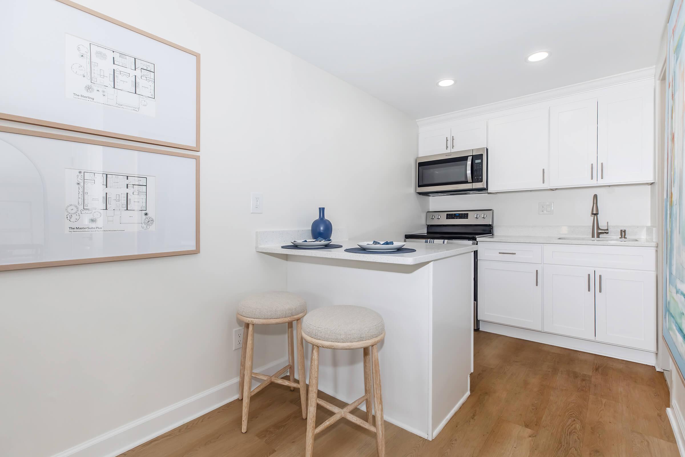 A modern kitchenette featuring white cabinetry, stainless steel appliances, and a compact dining area with two stools. Blue decorative elements are present, along with framed floor plans on the wall, enhancing the contemporary decor. Natural light brightens the space, highlighting the wood flooring.