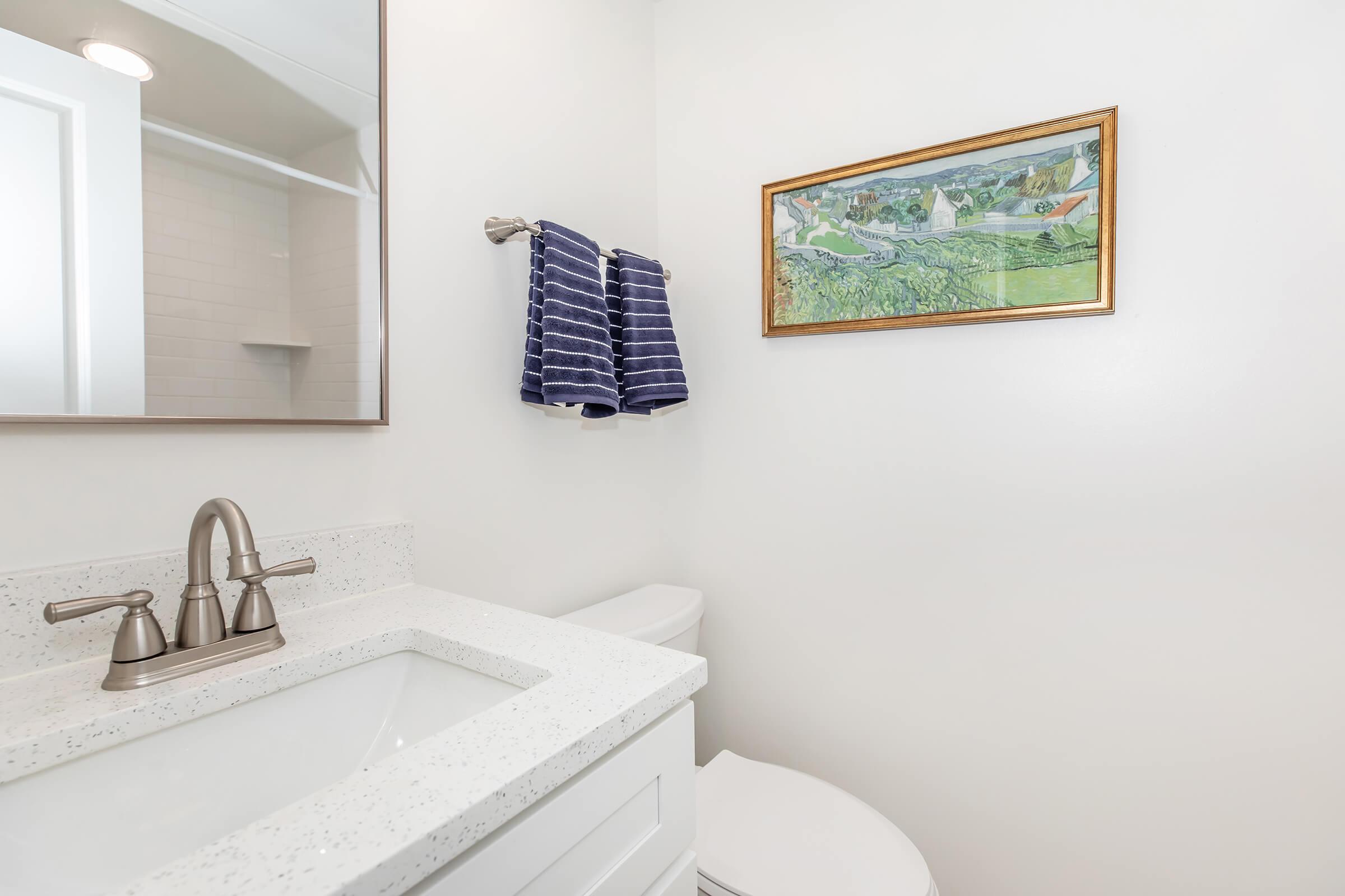 A modern bathroom featuring a white countertop sink, a mirror above, and a white toilet. A towel rack holds dark blue striped towels. On the wall, there is a framed painting of a countryside scene, adding an artistic touch to the minimalist design. The walls are painted a light color, enhancing the space's brightness.