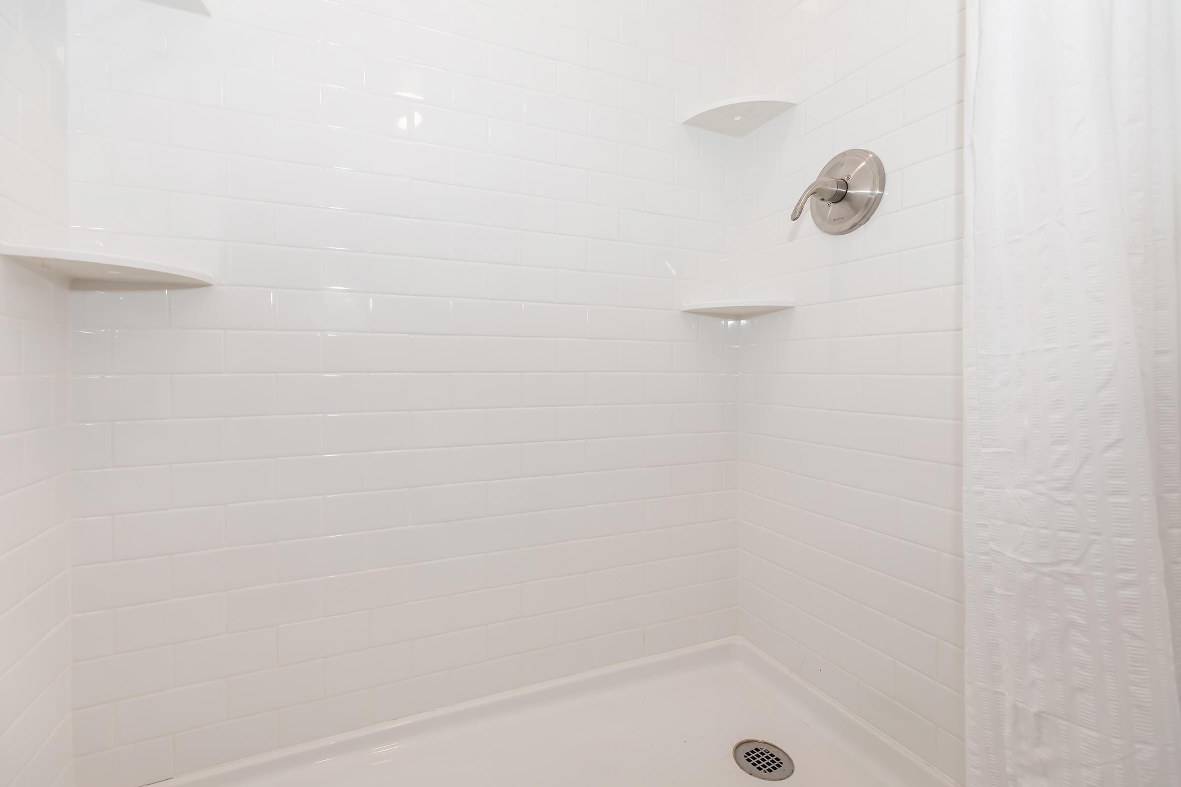 A clean, minimalist shower area featuring white tiled walls, three built-in corner shelves, a silver showerhead, and a white curtain. The floor is a simple shower base with a drain visible. The overall aesthetic is bright and modern.