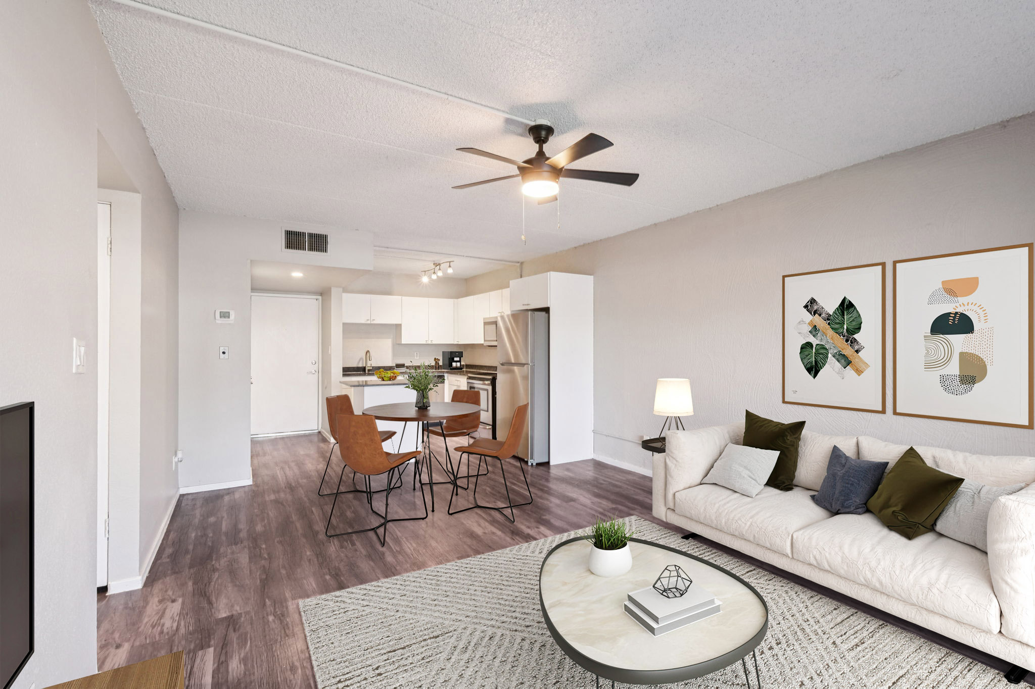 A modern living room featuring a light-colored sofa with decorative pillows, a round coffee table with a geometric centerpiece, and a ceiling fan. In the background, there is a dining area with a round table and four chairs, as well as a bright kitchen with contemporary appliances. Wall art adds color to the space.