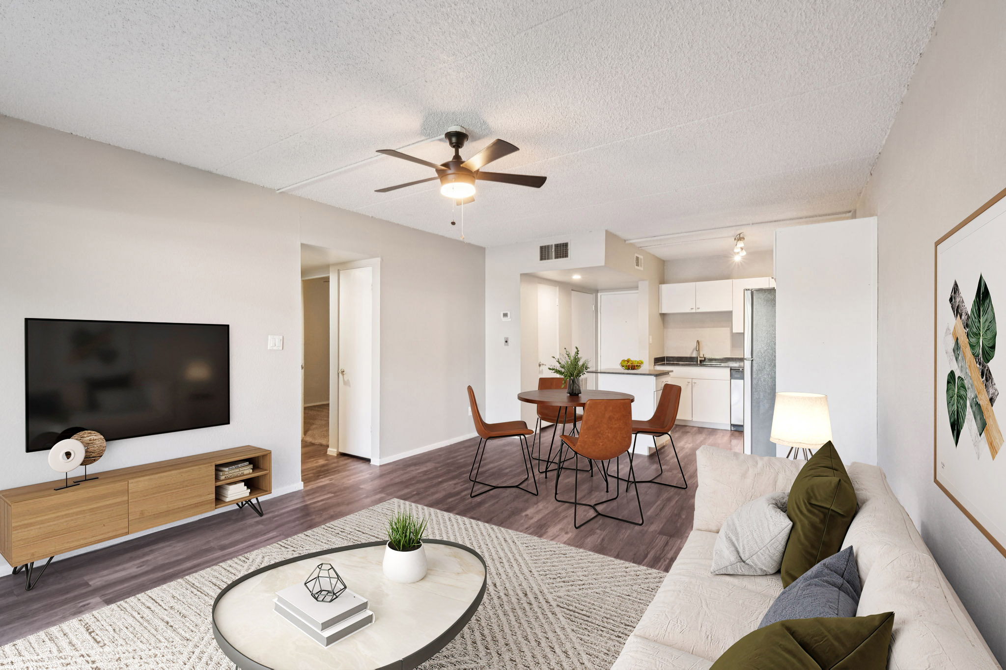 A modern living room featuring a light-colored sofa, a round coffee table with a plant centerpiece, and a television mounted on a wooden stand. In the background, a dining area with a round table and four chairs is visible, along with a glimpse of a kitchen. Bright, airy atmosphere with neutral tones.
