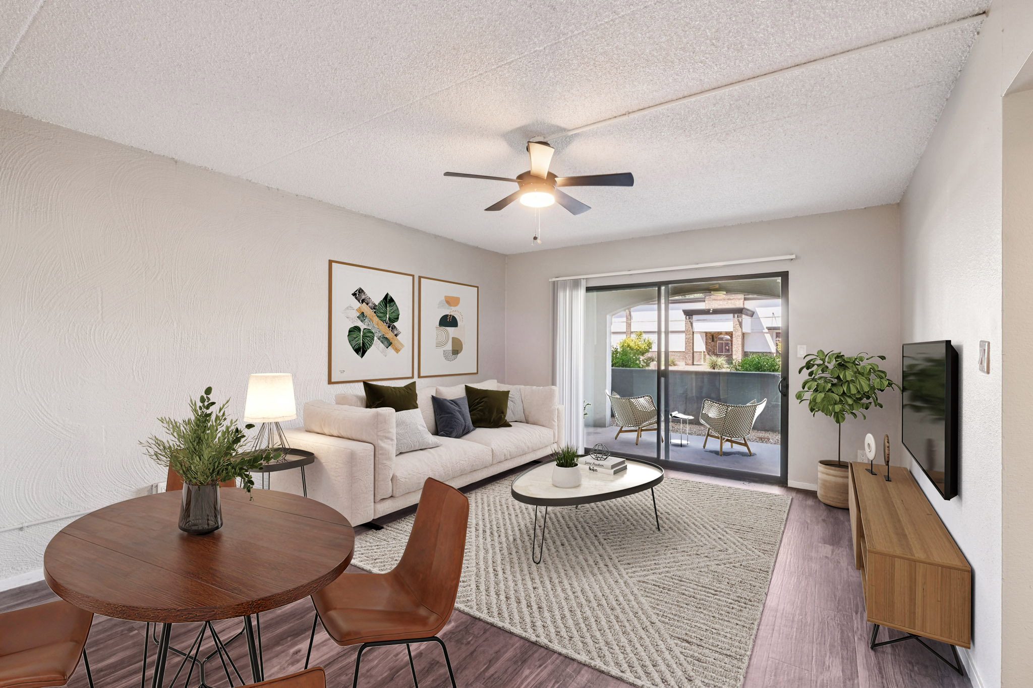 A modern living room featuring a comfortable white sofa with green and beige cushions, a round dining table with four brown chairs, and a decorative rug. A ceiling fan is visible, and there's a large window with a view of a balcony. Plants add a touch of greenery to the space.
