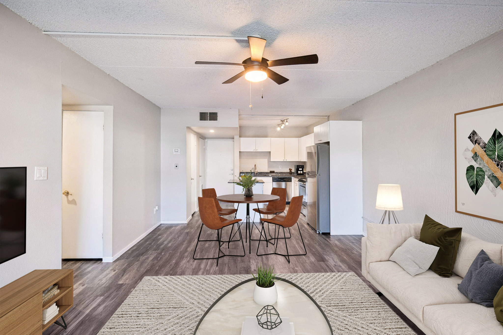 A modern living room with a ceiling fan, featuring a beige sectional sofa, a coffee table with a plant, and a TV unit. A dining area with four wooden chairs surrounds a round table. The kitchen is visible in the background, with white cabinets and appliances. Natural light brightens the space.