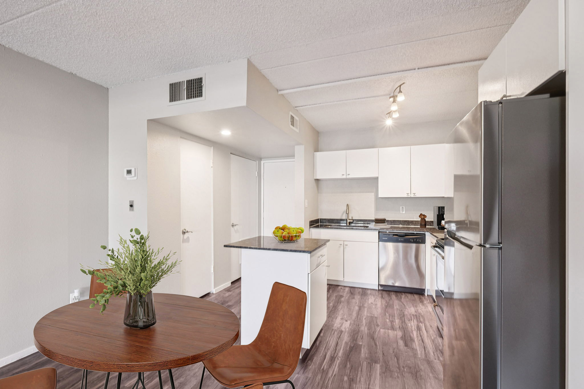 Modern kitchen and dining area with light gray walls, featuring white cabinets, stainless steel appliances, and a dark countertop. A round wooden table with four brown chairs is positioned near the kitchen, complemented by a vase of greenery. The space is well-lit and showcases an open layout.