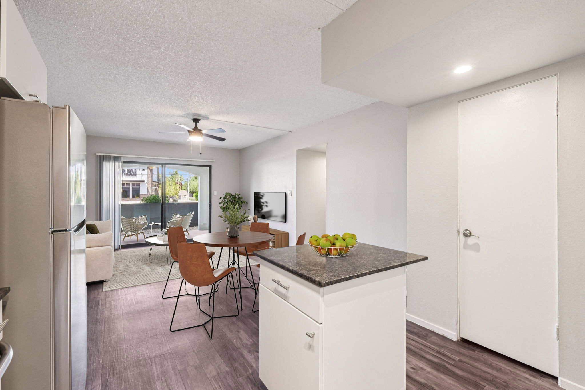 A modern open-concept kitchen and living area featuring a dining table with chairs, a kitchen island, a fridge, and a sleek TV area. The space has natural light coming through large windows, showcasing a cozy atmosphere with a bowl of apples on the kitchen island and a light color palette.