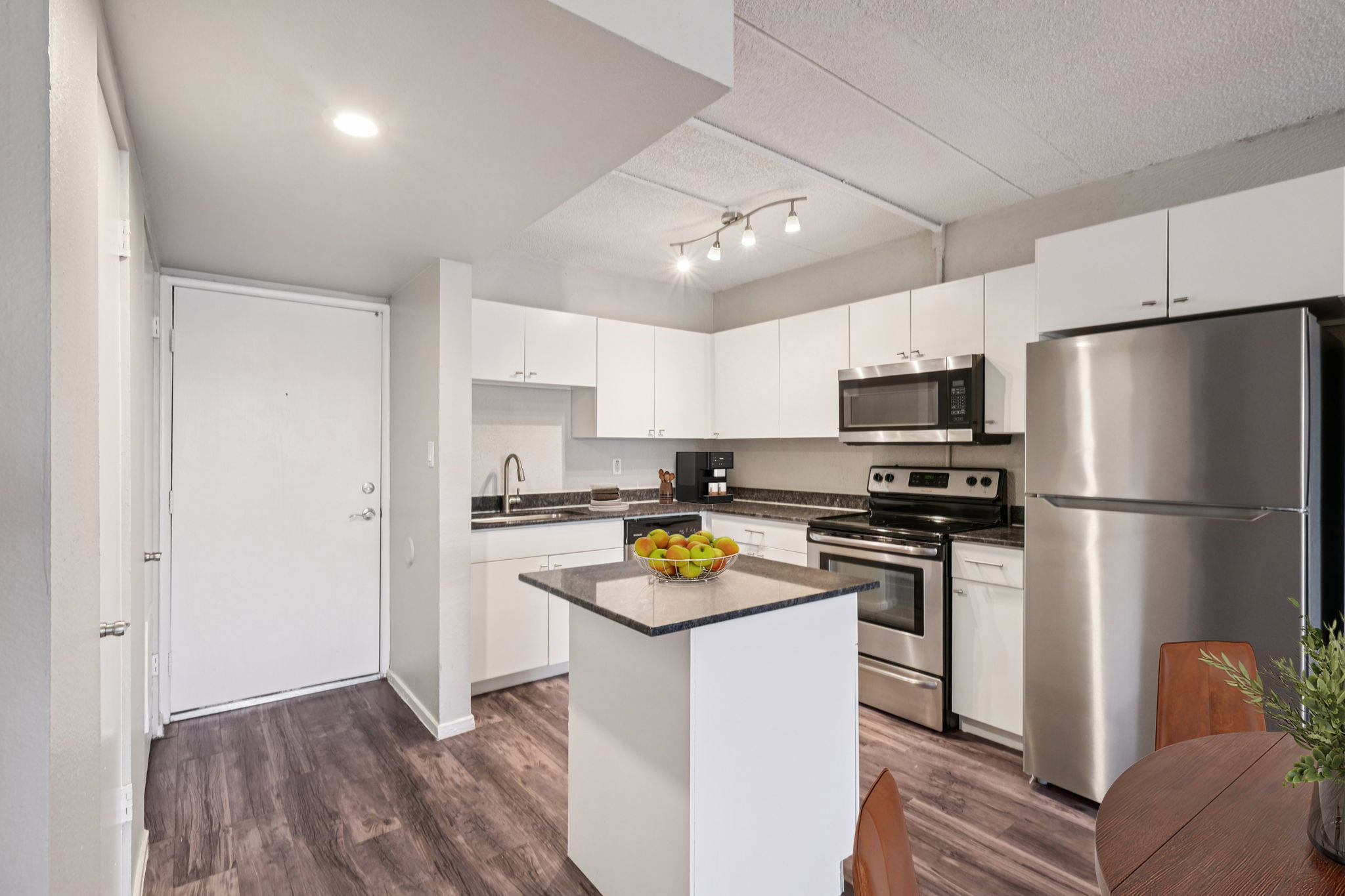 Modern kitchen with white cabinets, stainless steel appliances, and a dark countertop. Features include a silver refrigerator, microwave, and stove. A bowl of colorful fruit sits on the center island, and natural light illuminates the space, creating a welcoming atmosphere.