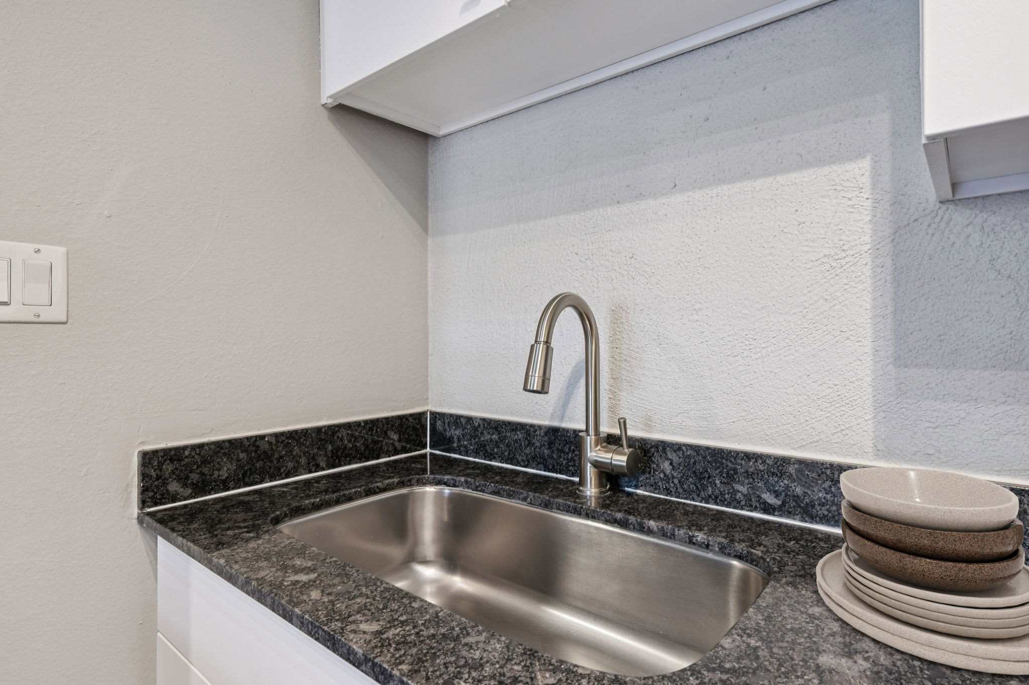 A modern kitchen sink with a stainless steel faucet on a dark granite countertop. Above the sink, there are white cabinets, and next to it, a stack of beige plates. The wall behind the sink is a light gray, creating a clean and contemporary look.