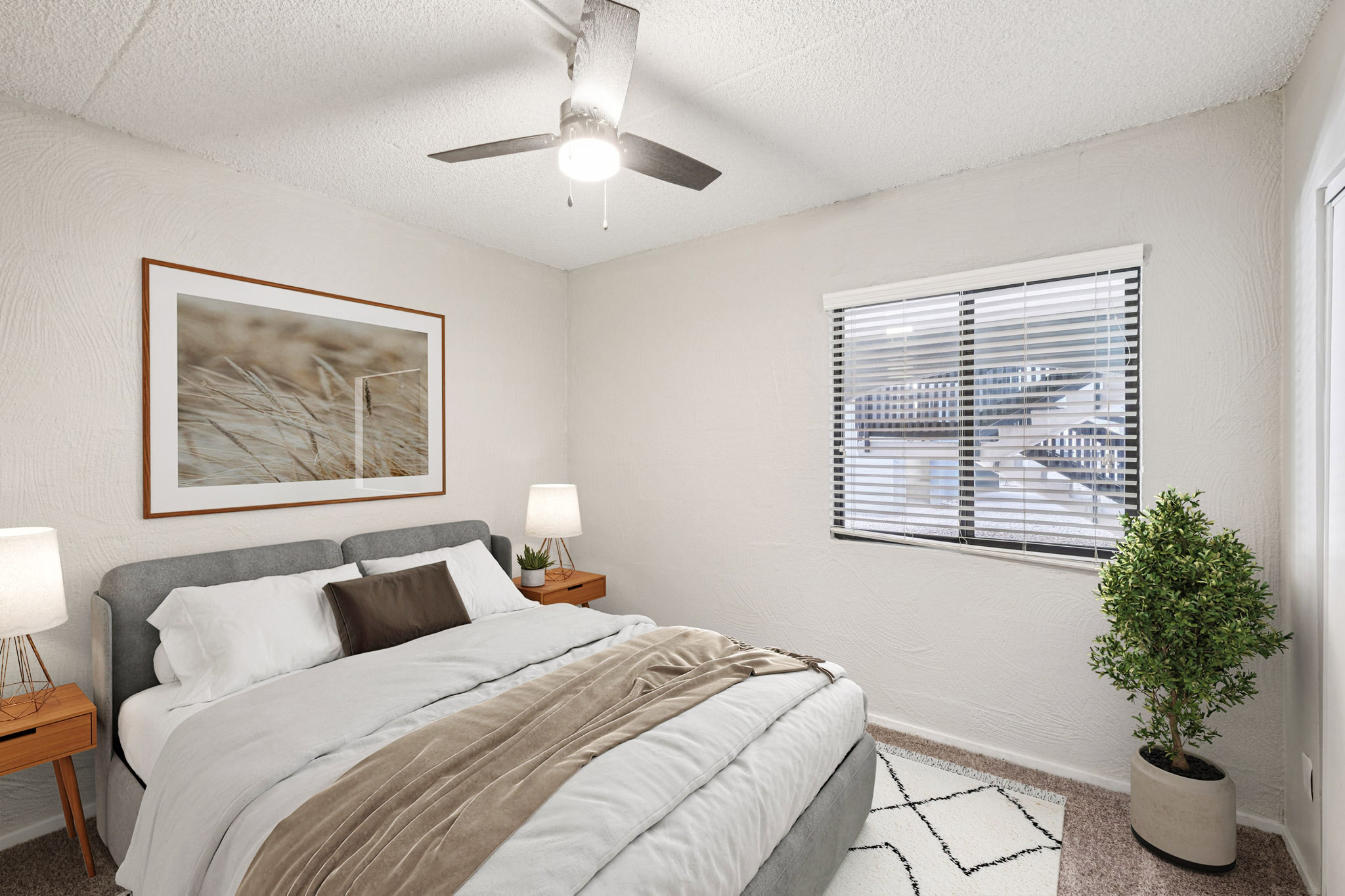 A cozy bedroom featuring a gray bed with white and brown bedding, two bedside tables with lamps, a potted plant, and a large framed photograph of grass on the wall. The room has light-colored walls, a ceiling fan, and a window with horizontal blinds allowing natural light to enter.