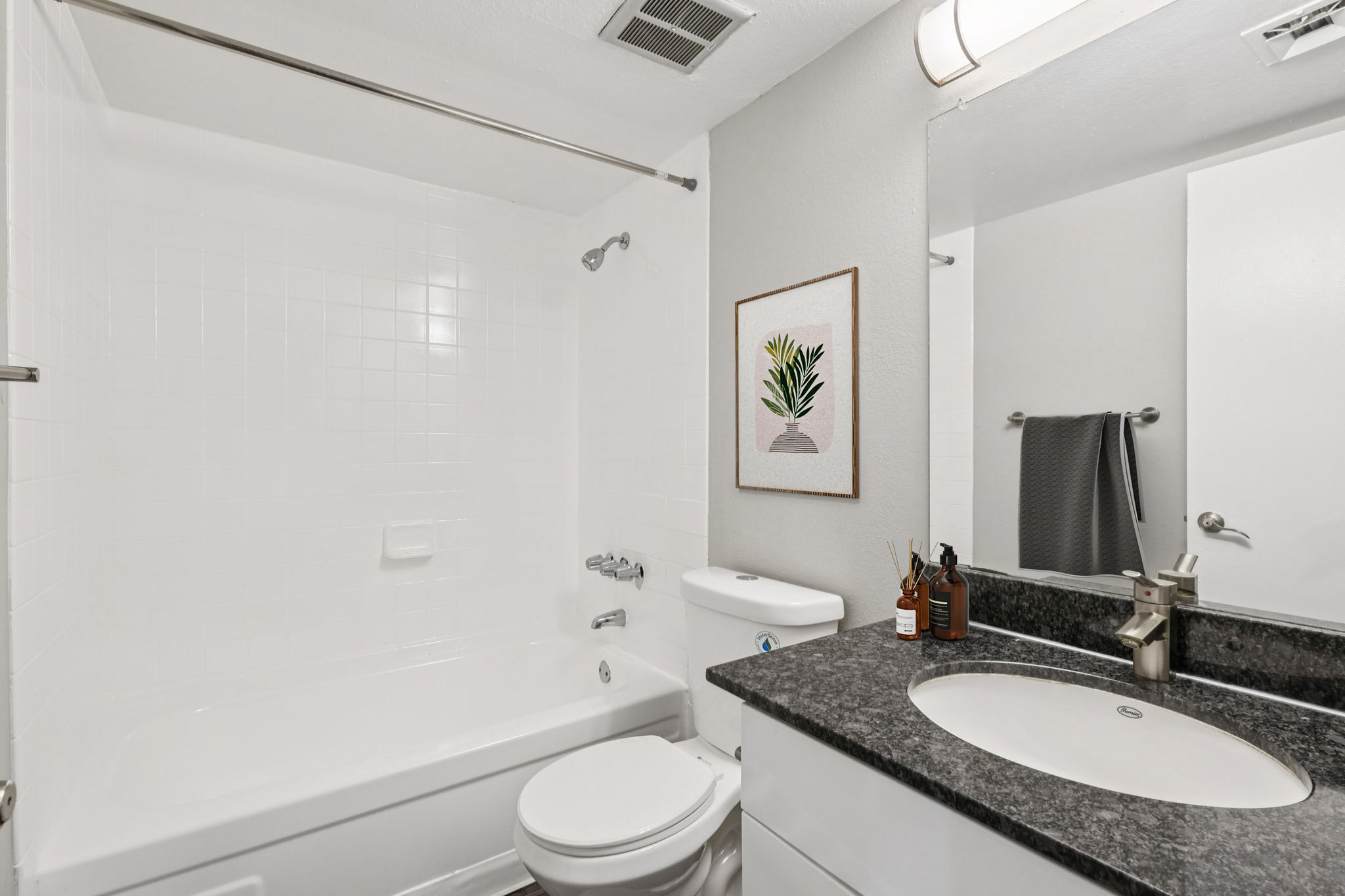 A clean bathroom featuring a white tiled shower and bathtub combination, a modern toilet, and a vanity with a dark countertop. A mirror above the sink reflects the space, and a framed artwork of a plant hangs on the wall. Towels are neatly displayed, enhancing the overall aesthetic.