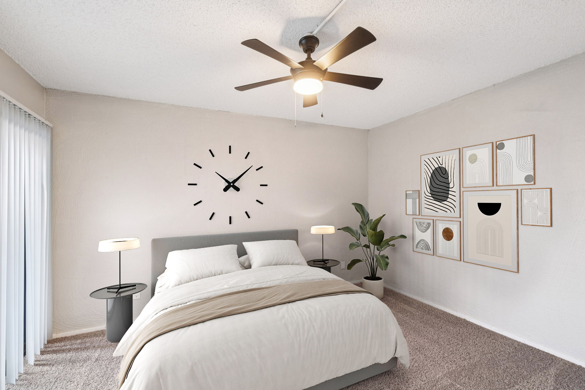 A modern bedroom featuring a gray bed with white bedding, two nightstands with table lamps, a wall clock above the bed, and a collection of framed wall art. There is a leafy plant in the corner and sliding glass doors allowing natural light to brighten the space. The room has a neutral color palette.