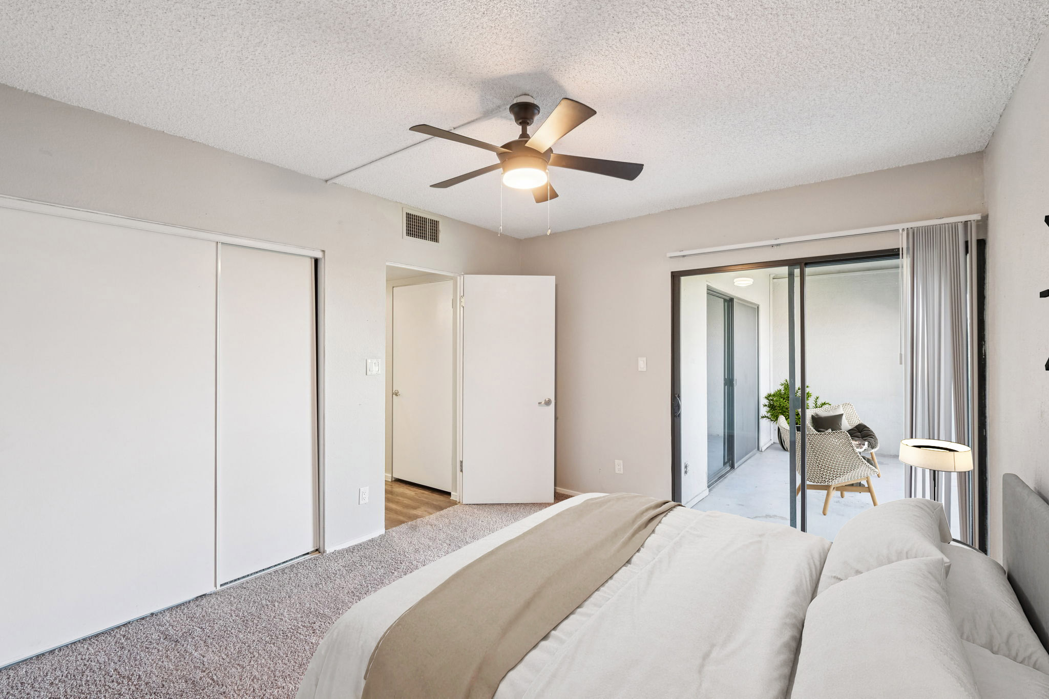 A well-lit bedroom featuring a ceiling fan, light-colored walls, and carpeting. There are closet doors on one side and a sliding glass door leading to a patio area. The bed is made with neutral-colored bedding, and a touch of greenery is visible through the patio door.