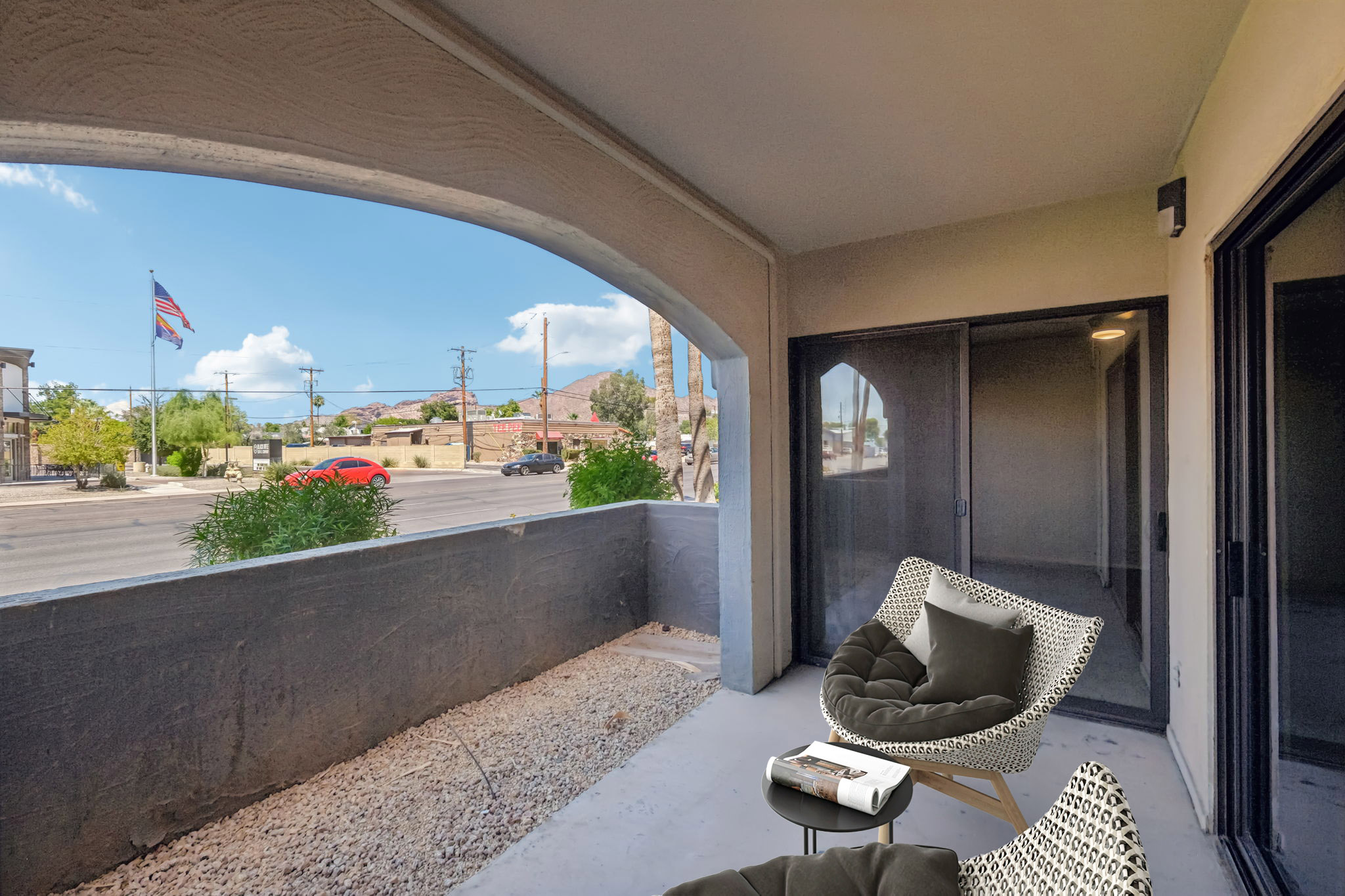 A cozy balcony view featuring two modern chairs, with gravel flooring and a black railing. In the background, an American flag is visible along with a street scene that includes trees, cars, and distant buildings under a blue sky with a few clouds.