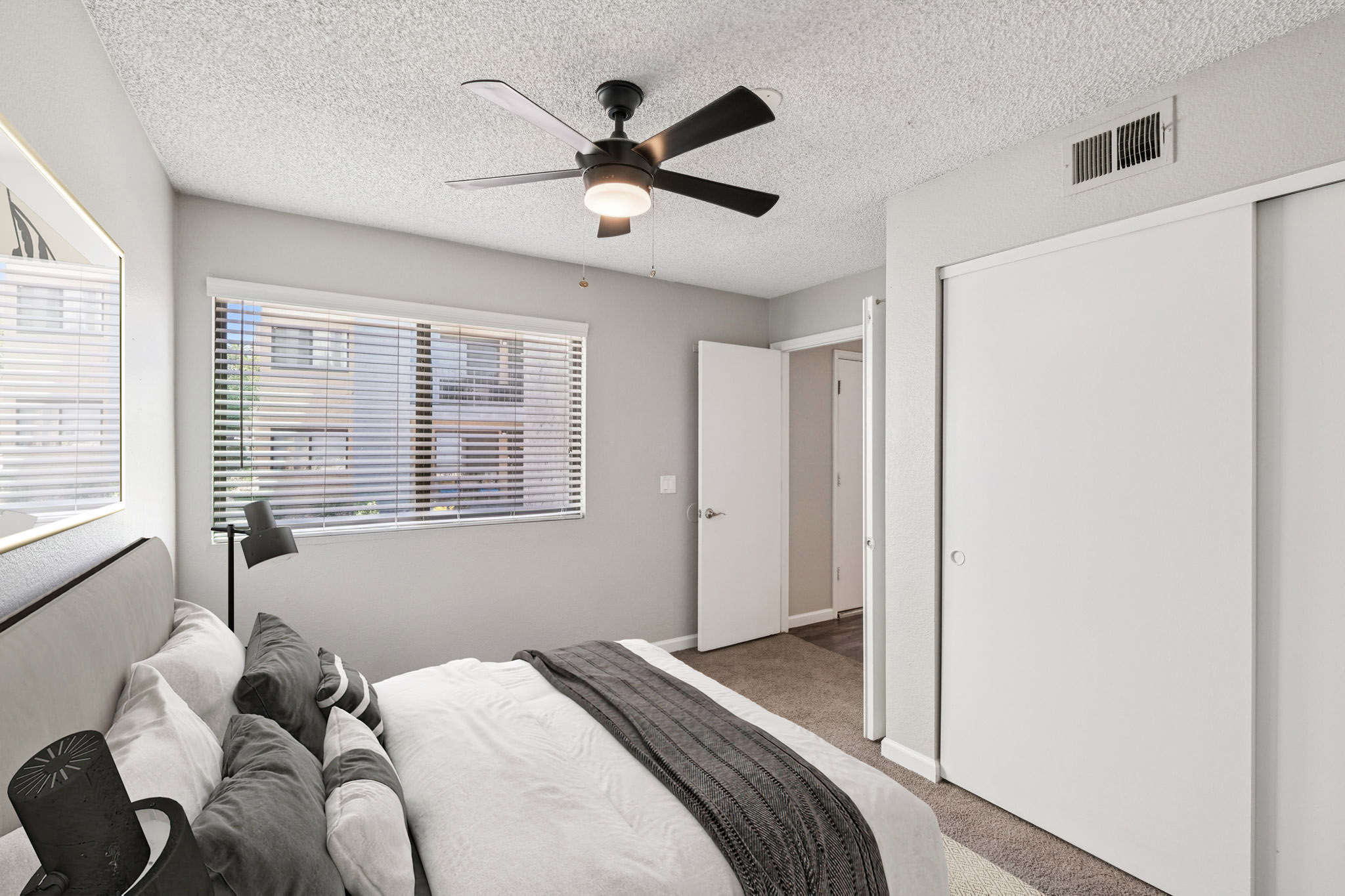 A modern bedroom featuring a gray color scheme. The room includes a neatly made bed with gray and white bedding, a ceiling fan, and a bedside lamp. A window with blinds allows natural light to enter, and there is a door leading to another room, along with a closet. The overall atmosphere is calm and inviting.