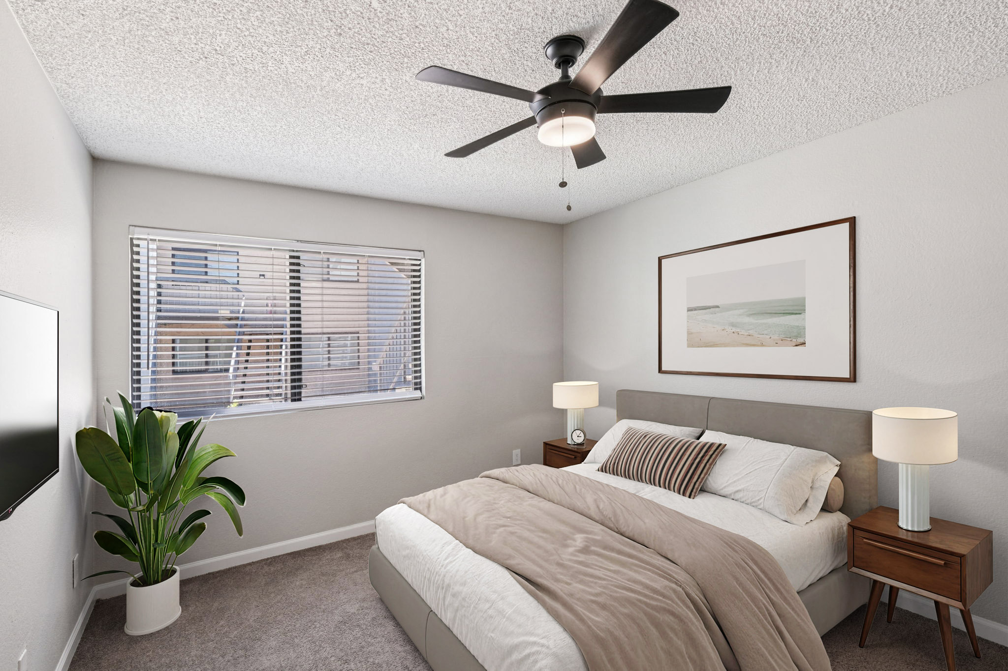 A cozy bedroom featuring a comfortable bed with a plush beige blanket, two bedside tables with lamps, a ceiling fan, and a large window with blinds. A potted plant adds a touch of greenery, while a framed beach photograph decorates the wall, creating a serene and inviting atmosphere.