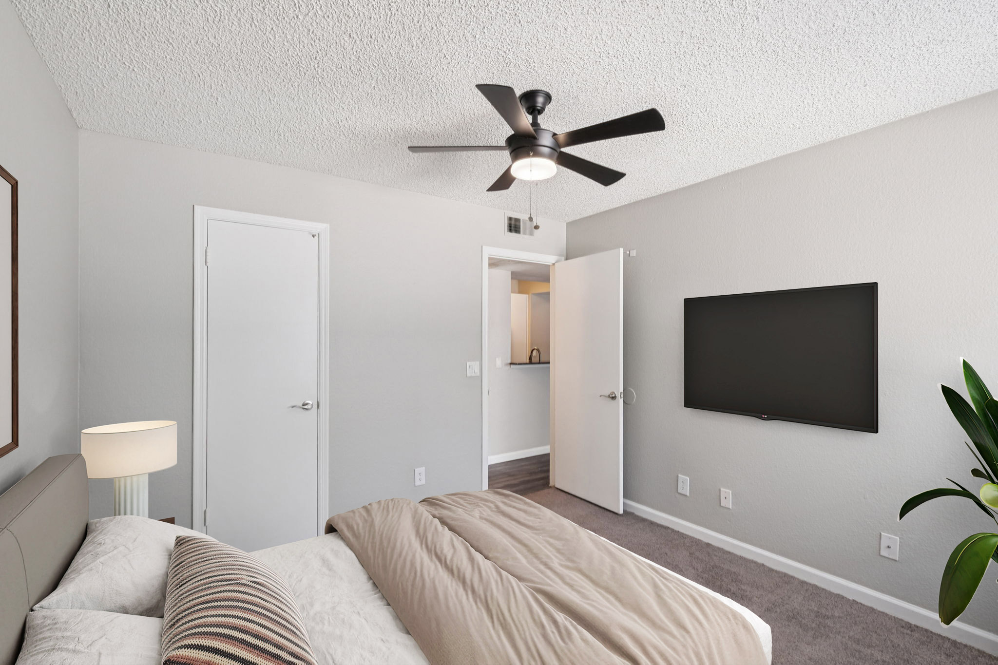 A cozy bedroom featuring a ceiling fan, a large wall-mounted TV, and a neatly made bed with a light-colored blanket. The walls are painted in a soft gray tone, and there's a small nightstand with a lamp beside the bed. An open door leads to a kitchenette, and a potted plant adds a touch of greenery.