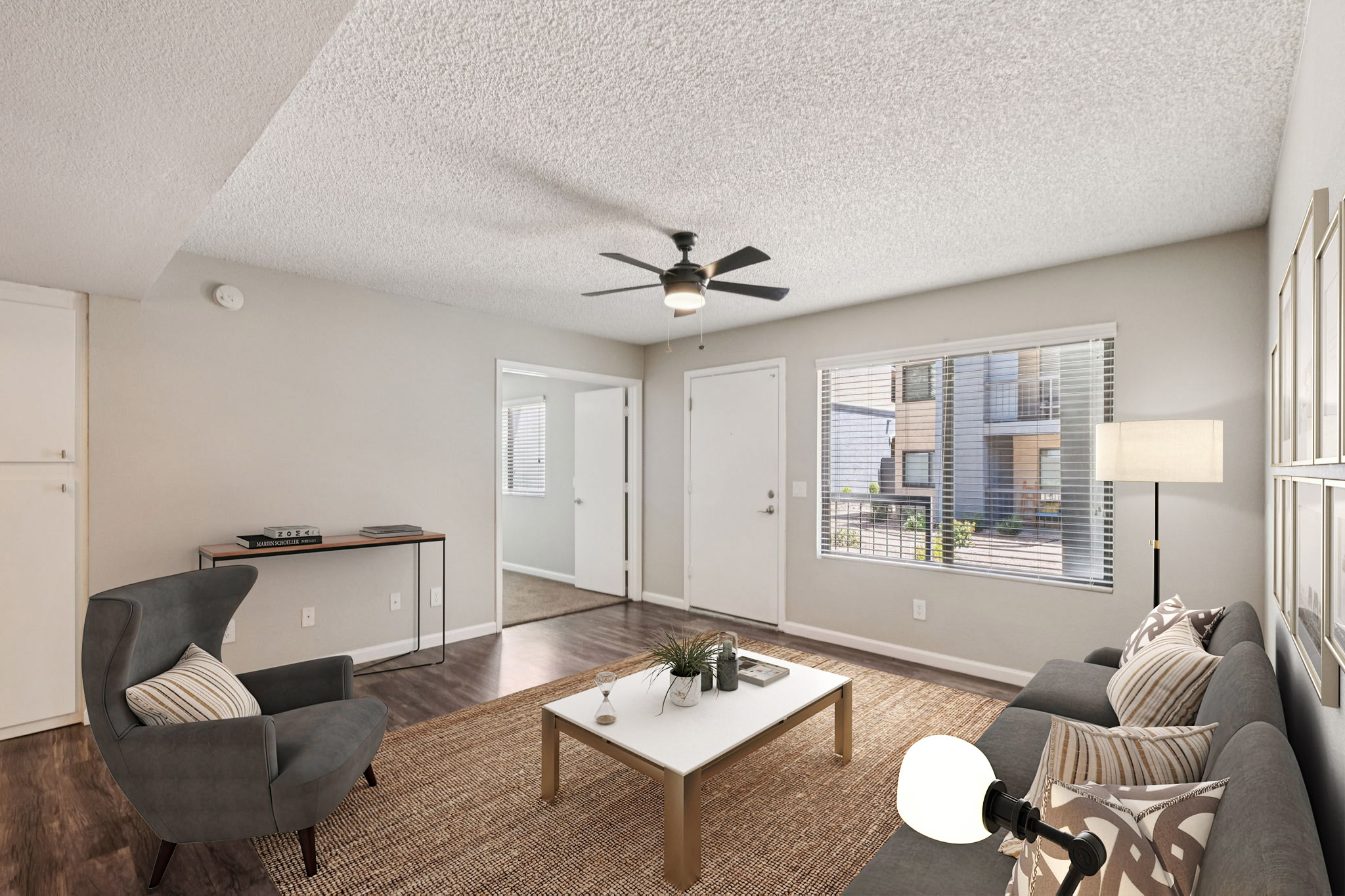 A cozy living room featuring gray sofas with decorative pillows, a coffee table with a plant, and a light fixture. The space has a ceiling fan, large windows that allow natural light, and a neutral color palette. Wood flooring adds warmth to the atmosphere.
