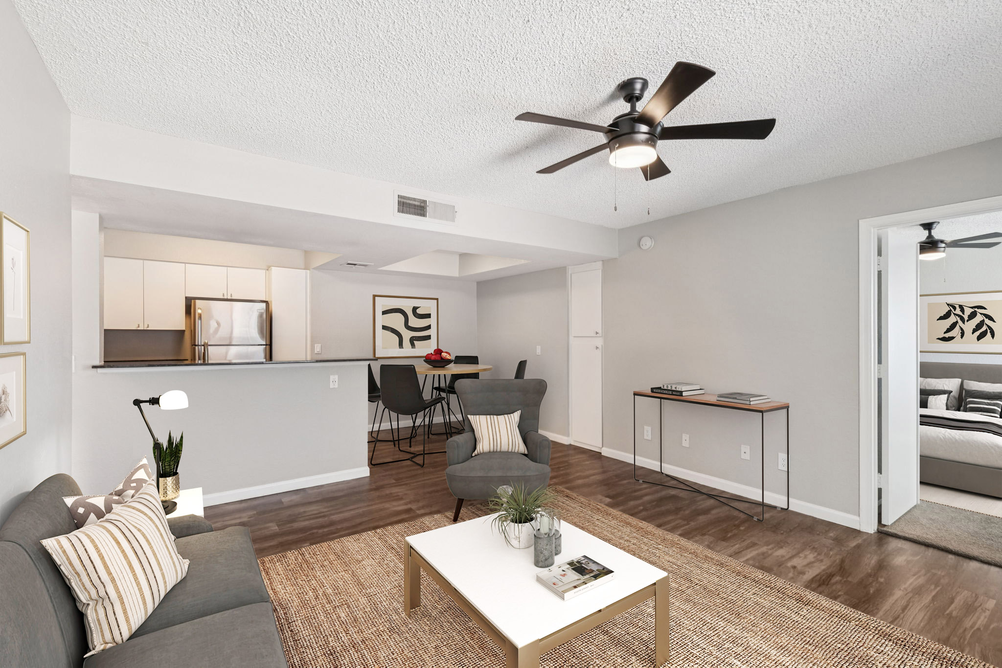 A stylish living room featuring a gray sofa with decorative pillows, a white coffee table with a plant, and a ceiling fan. The space opens to a kitchen area with light cabinets and stainless steel appliances. A dining table with chairs is visible, along with a doorway leading to a bedroom.
