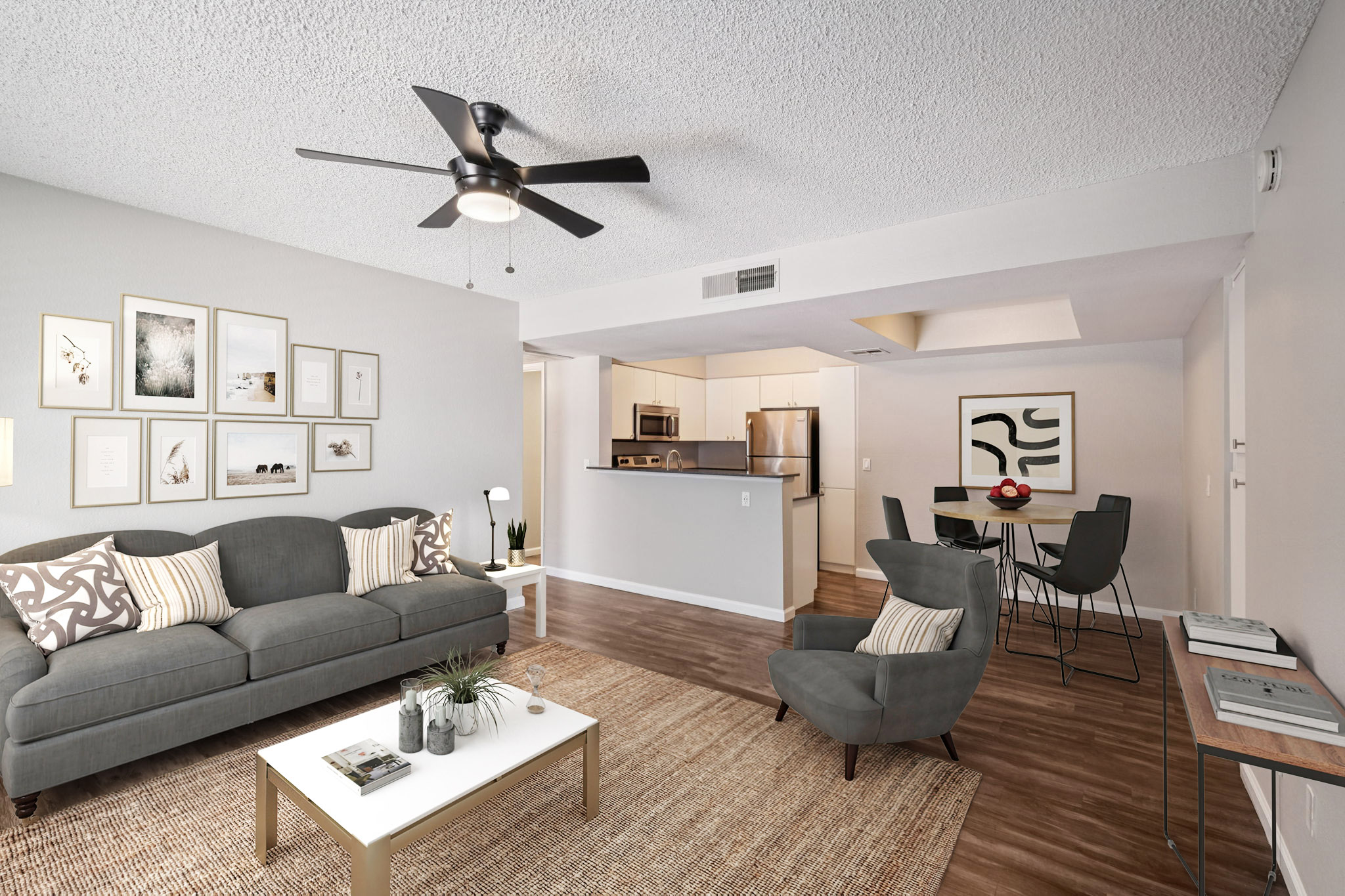 A modern living room featuring a gray sofa with decorative pillows, a stylish coffee table, and an area rug. A ceiling fan is overhead, and framed artwork adorns the walls. In the background, there is a dining area with a small table and chairs, adjacent to a kitchen with stainless steel appliances.