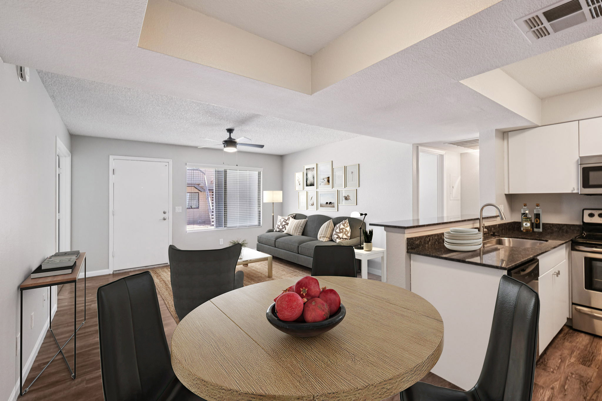 Modern living space featuring an open-plan design. A round dining table with a bowl of red apples is at the center, surrounded by black chairs. In the background, a cozy living area with a gray sofa and decorative wall art can be seen, along with a kitchen equipped with stainless steel appliances. Natural light enters through a window.
