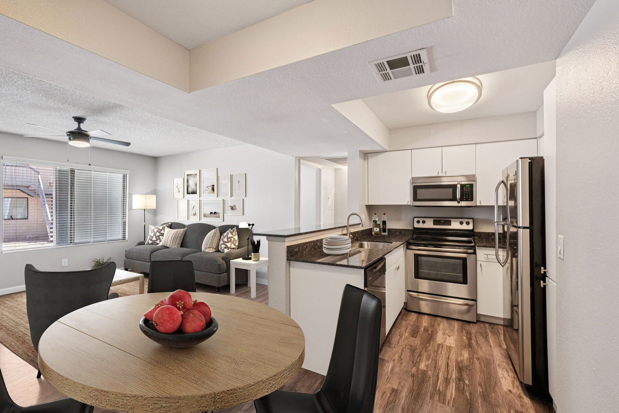 Modern kitchen and dining area in a well-lit living space. The kitchen features stainless steel appliances, a sink, and ample counter space. A round wooden dining table with a bowl of red fruit sits nearby. The living area has a cozy gray sofa and decorative wall art. Wood-style flooring enhances the contemporary look.