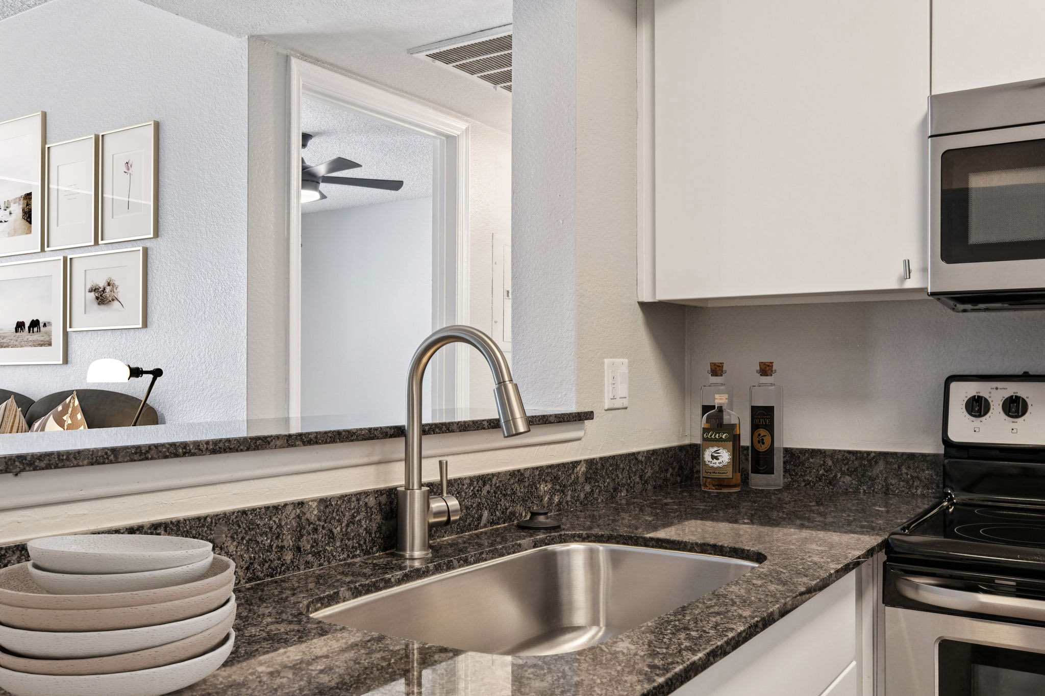 A modern kitchen featuring gray granite countertops, a stainless steel sink, and a sleek faucet. In the background, there are decorative items on the counter, including bottles. The kitchen connects to a light-filled living space with framed artwork on the walls and a fan overhead.