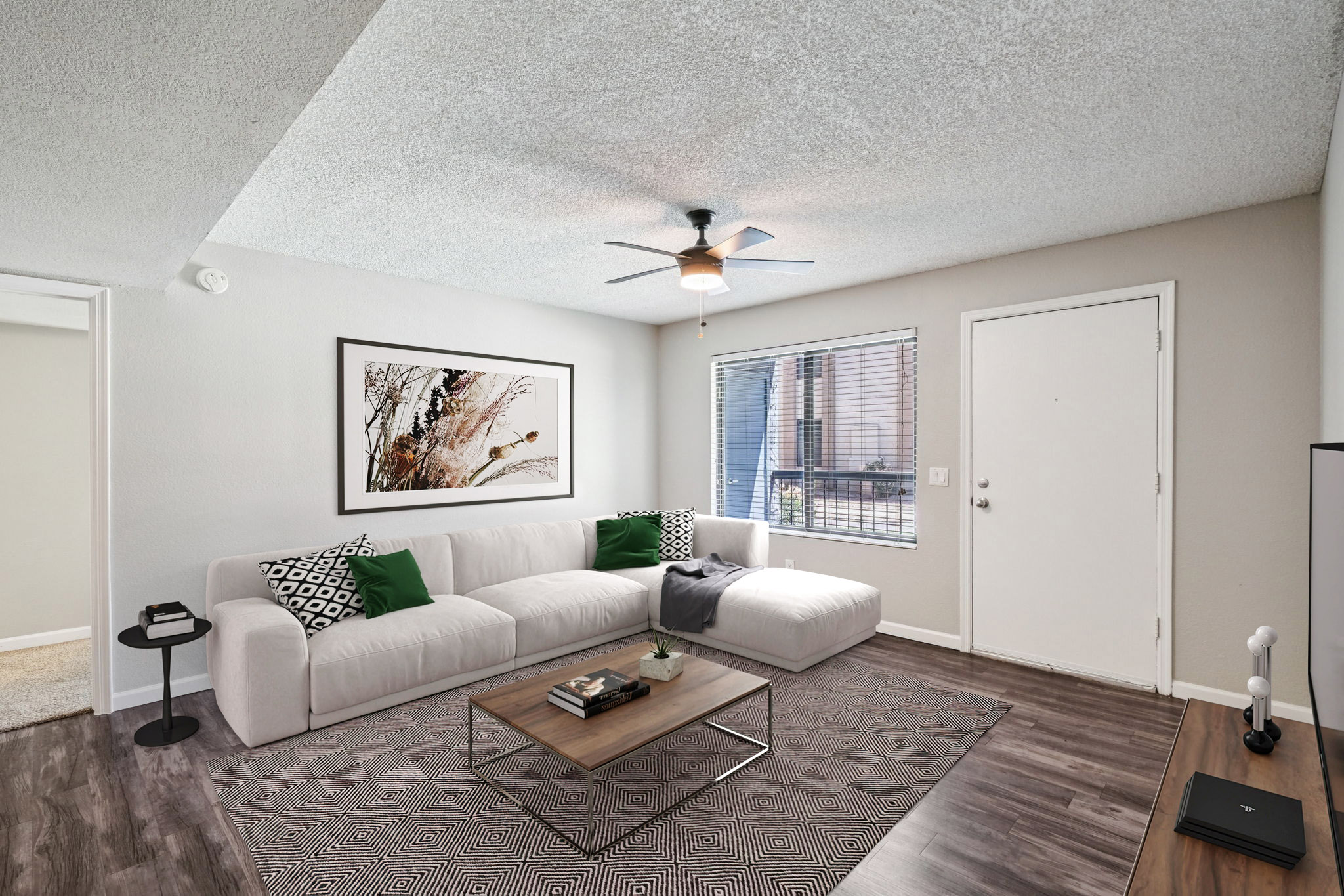 A modern living room featuring a white sectional sofa with green decorative pillows, a wooden coffee table, and a textured area rug. A ceiling fan hangs above, and large windows provide natural light. The walls are painted in neutral tones, and there’s an open door leading to an exterior space.