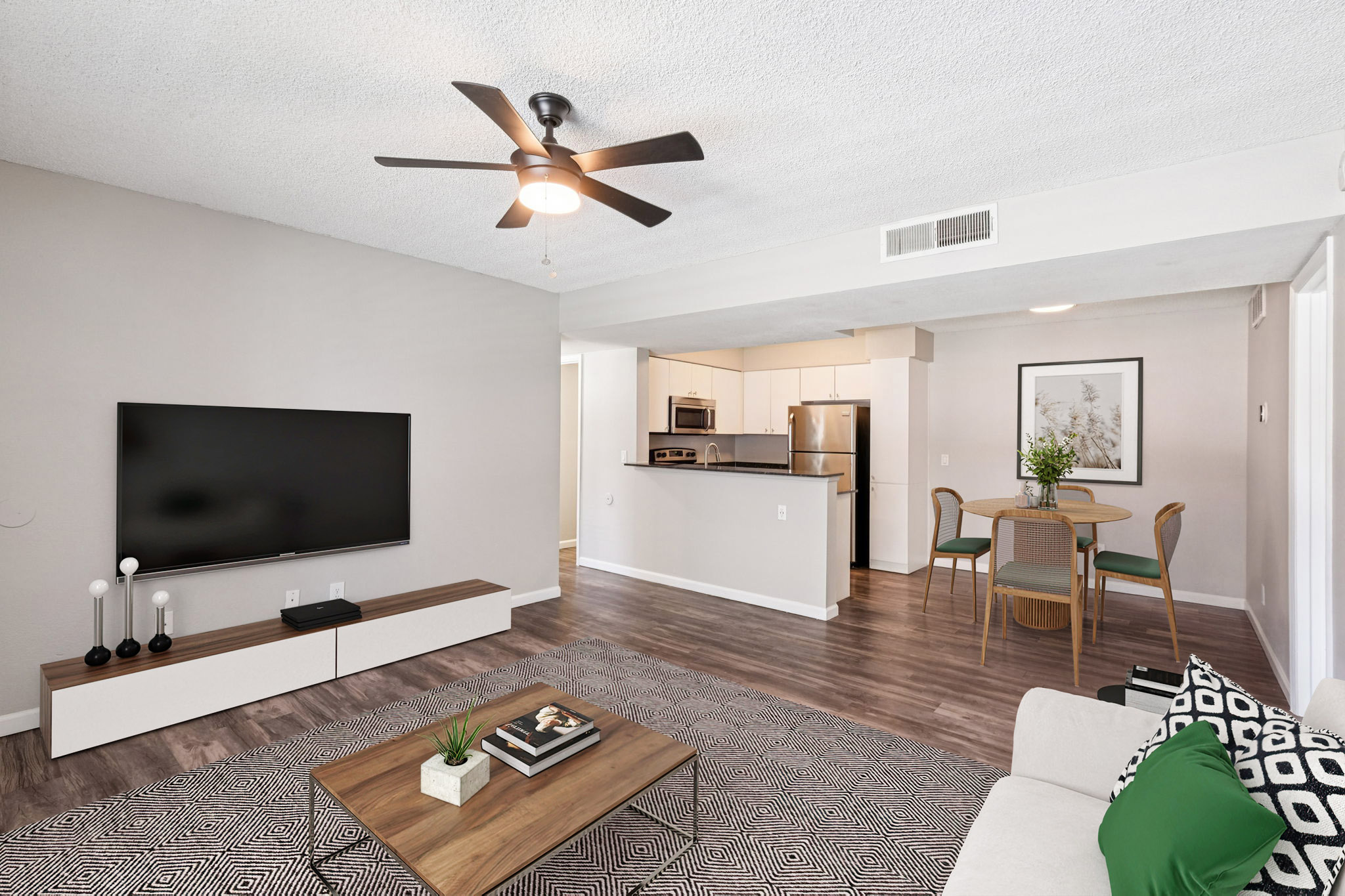 A modern living room featuring a light couch, coffee table, and a TV mounted on the wall. A dining area is visible in the background with a round table and chairs. Ceiling fan above provides a cozy atmosphere. Neutral color palette and wooden flooring create a warm, inviting space. Decorative plants and accessories complete the decor.