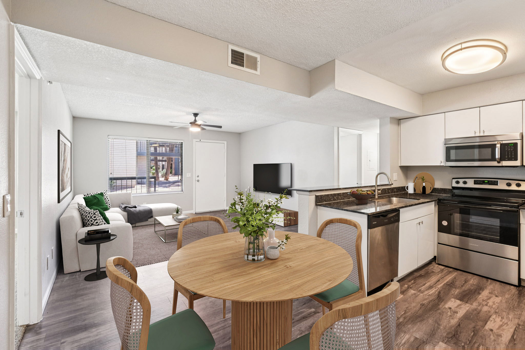 A modern apartment interior featuring a dining area with a round wooden table and chairs, a kitchen with stainless steel appliances, and a living space with a couch and TV. Natural light flows through a window, and there are decorative plants and neutral-colored walls in a spacious layout.