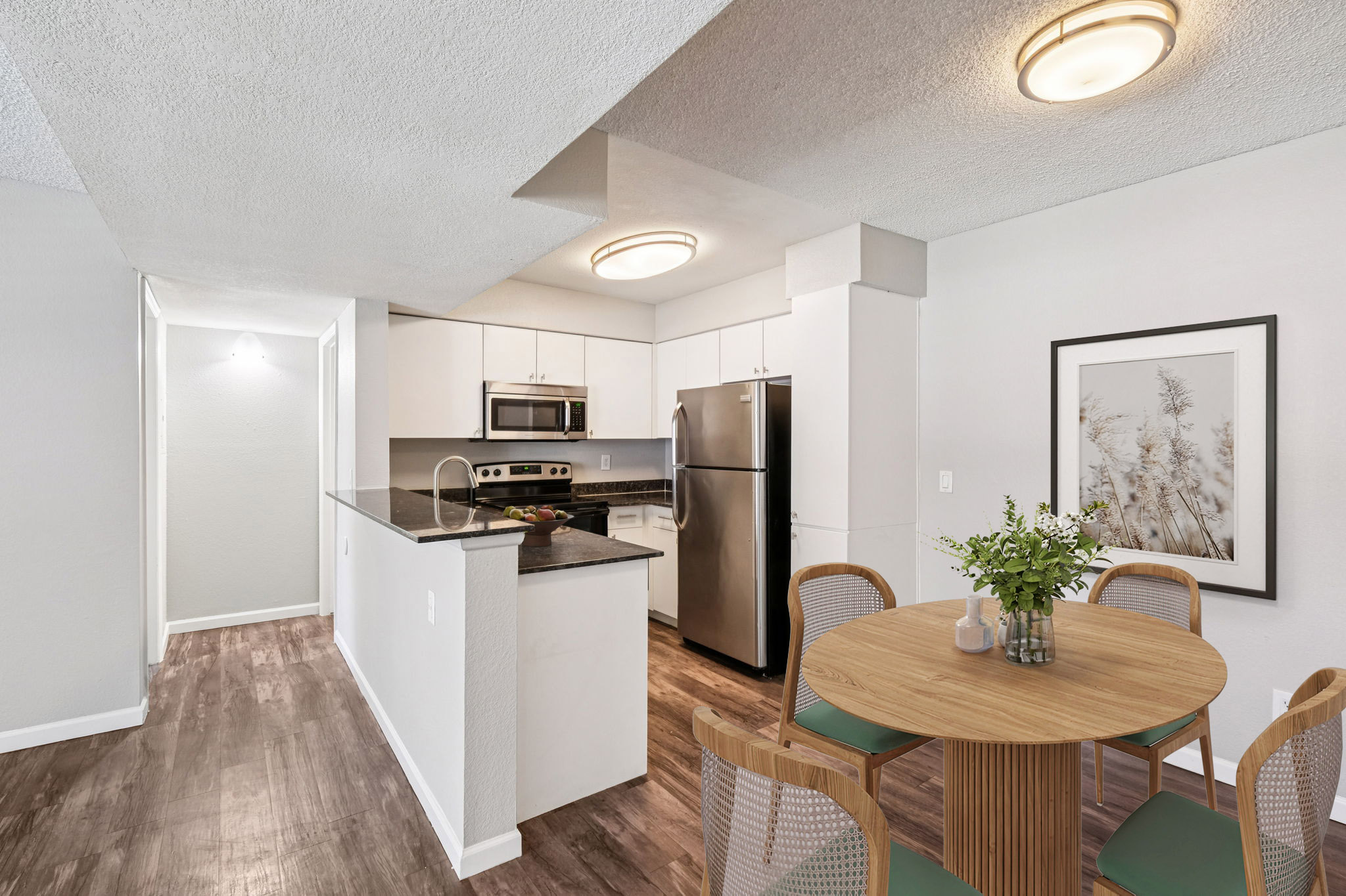 Modern kitchen and dining area featuring sleek white cabinetry, stainless steel appliances, and a round wooden dining table with stylish chairs. The space is well-lit with overhead lighting and has a neutral color palette, creating a fresh and inviting atmosphere.