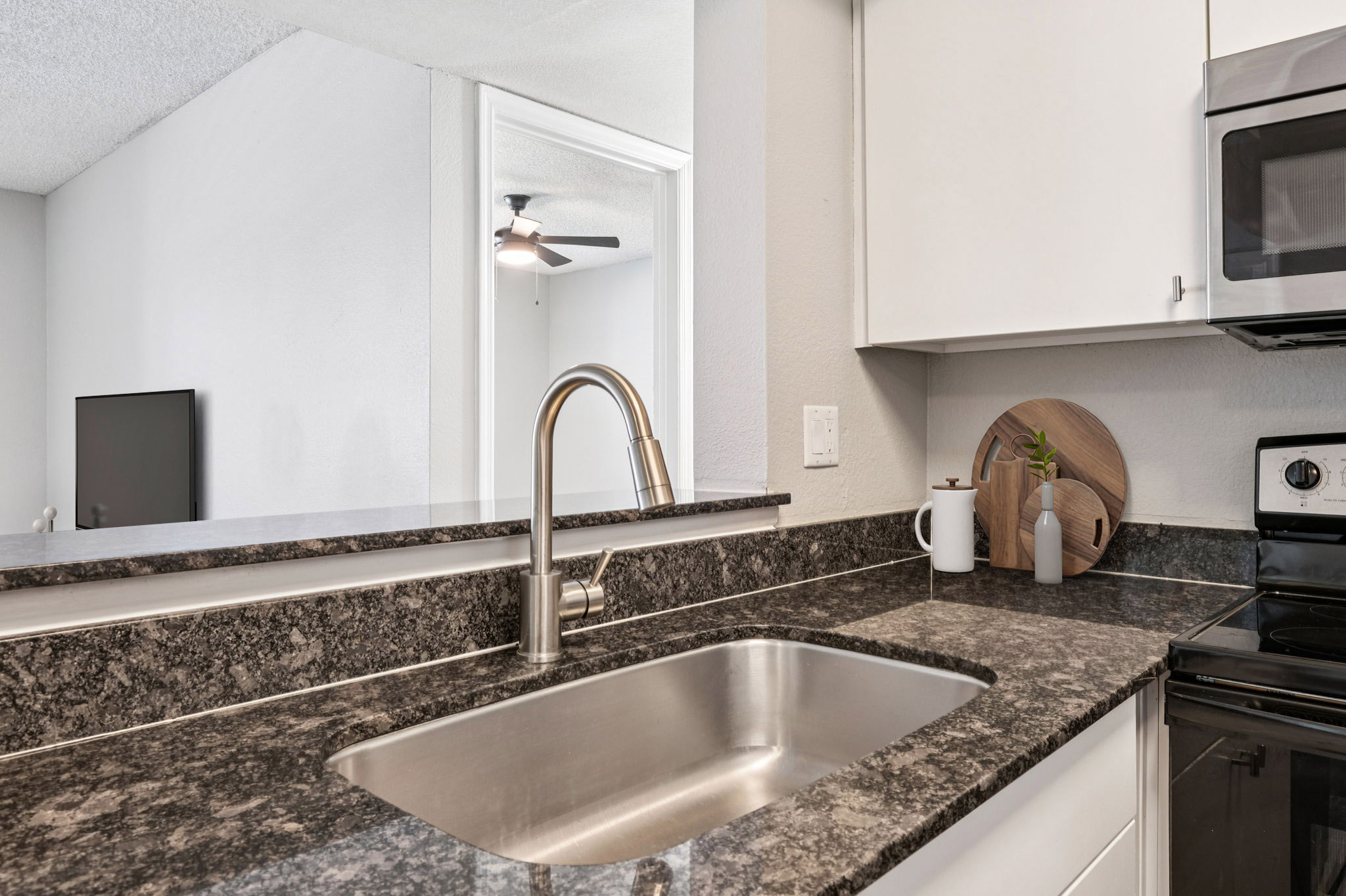 A modern kitchen with a stainless steel sink, dark granite countertop, and white cabinetry. In the background, a doorway leads to a well-lit living area with a ceiling fan and a TV. Stylish decor includes a wooden cutting board and a small plant on the countertop.