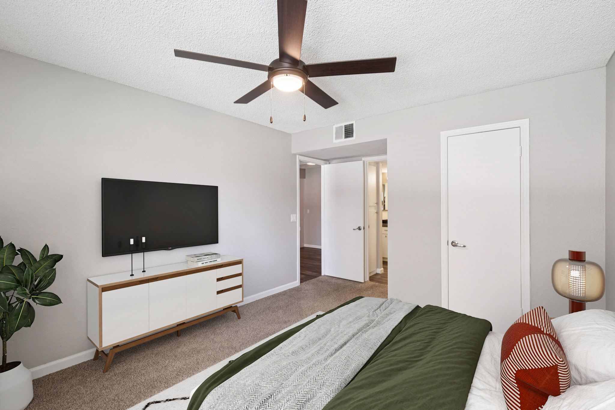 A modern bedroom featuring a bed with a green blanket, a flat-screen TV mounted on the wall, a stylish dresser, and a potted plant. The room has light-colored walls, a ceiling fan, and a door leading to a bathroom. Natural light enhances the cozy atmosphere with contemporary decor accents.