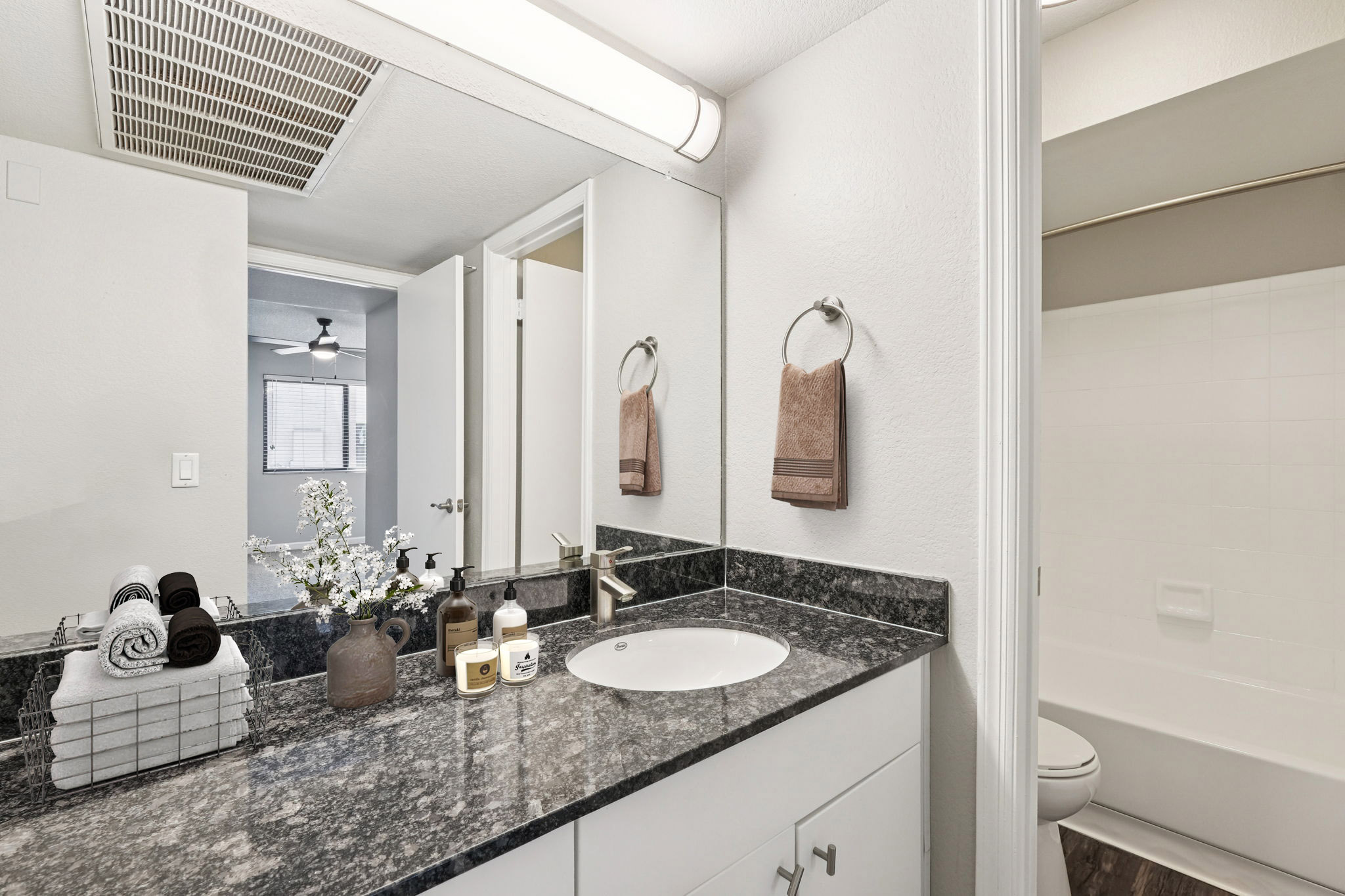 A modern bathroom featuring a granite countertop with a round sink, neatly arranged towels in a basket, and decorative toiletries. A shower area is visible in the background along with light-colored walls and a mirror above the sink. Natural light enters from a window, giving a spacious feel.