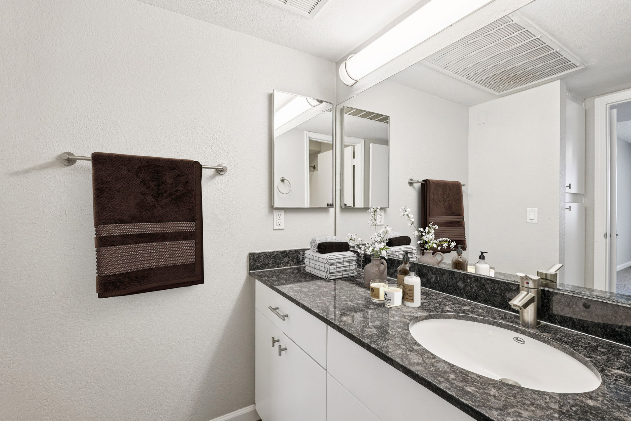 A modern bathroom featuring a gray countertop with a circular sink, two mirrors, and toiletries. White walls and a towel rack with dark brown towels are visible. The decor includes small plants and candles, enhancing the clean and minimalist aesthetic. A glimpse of a closet can be seen in the background.