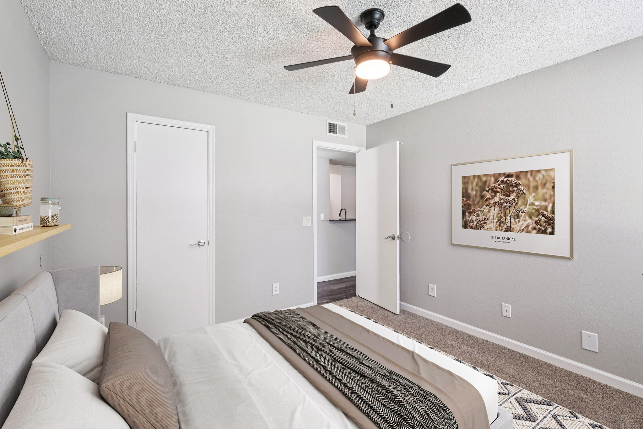 A cozy bedroom featuring a queen-sized bed with neutral bedding, a textured throw blanket, and a bedside lamp. The room has light gray walls, a ceiling fan, and a modern piece of framed artwork on the wall. A door leads to another room, adding to the spacious feel.