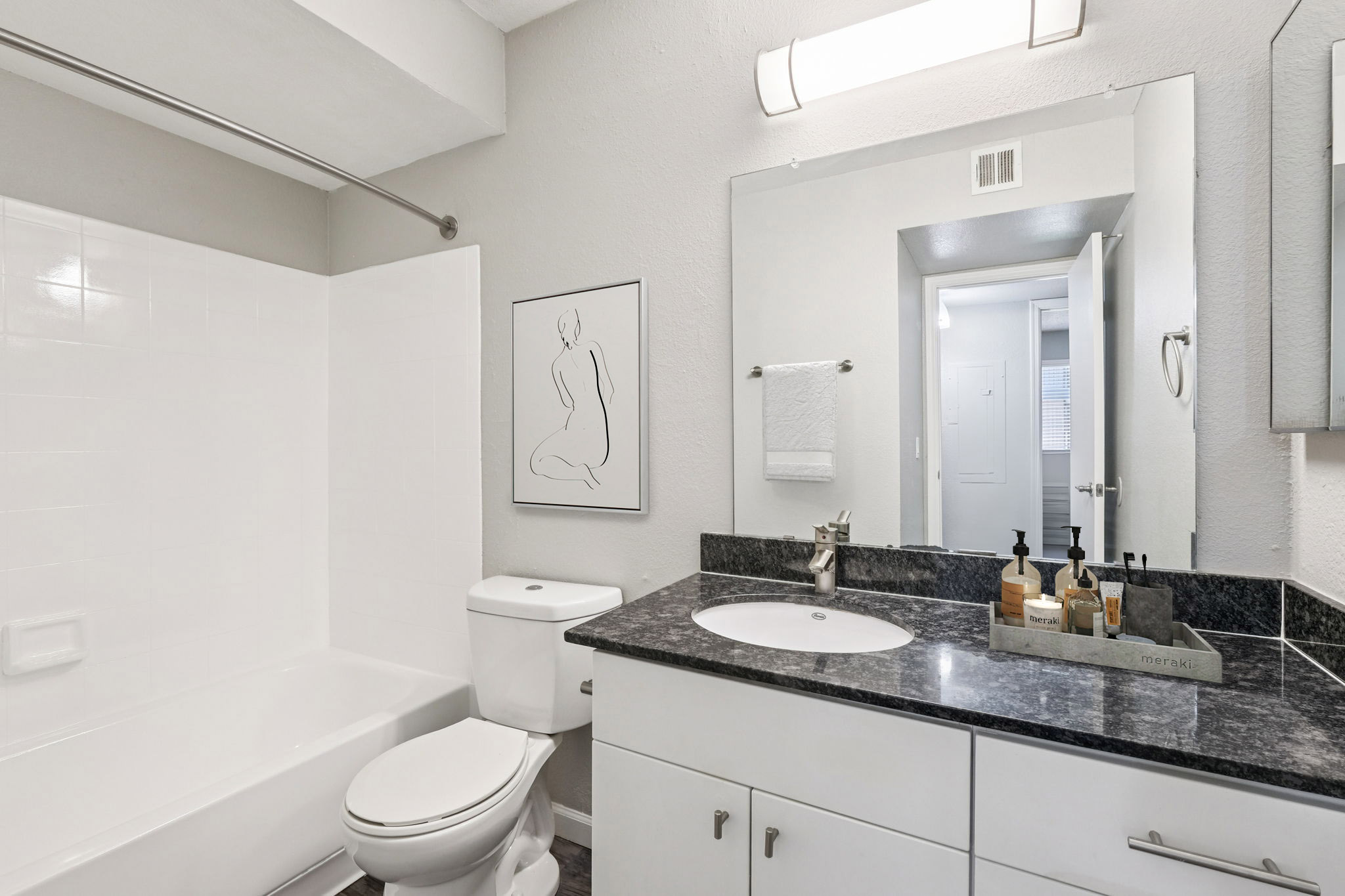 A modern bathroom featuring a white bathtub with a shower, a sleek sink with a dark granite countertop, a large mirror, and tasteful decor. There are toiletries neatly arranged on the countertop, a framed art piece on the wall, and light-colored walls that create a clean, minimalist aesthetic.