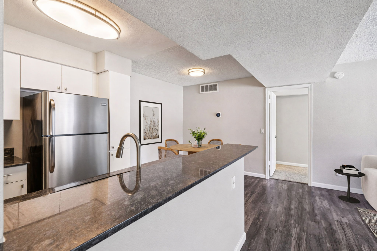 Modern kitchen and dining area featuring a stainless steel refrigerator, a sleek black countertop, and a wooden dining table with chairs. The walls are painted in neutral tones, and there’s a doorway leading to another room. Soft lighting fixtures enhance the inviting atmosphere.